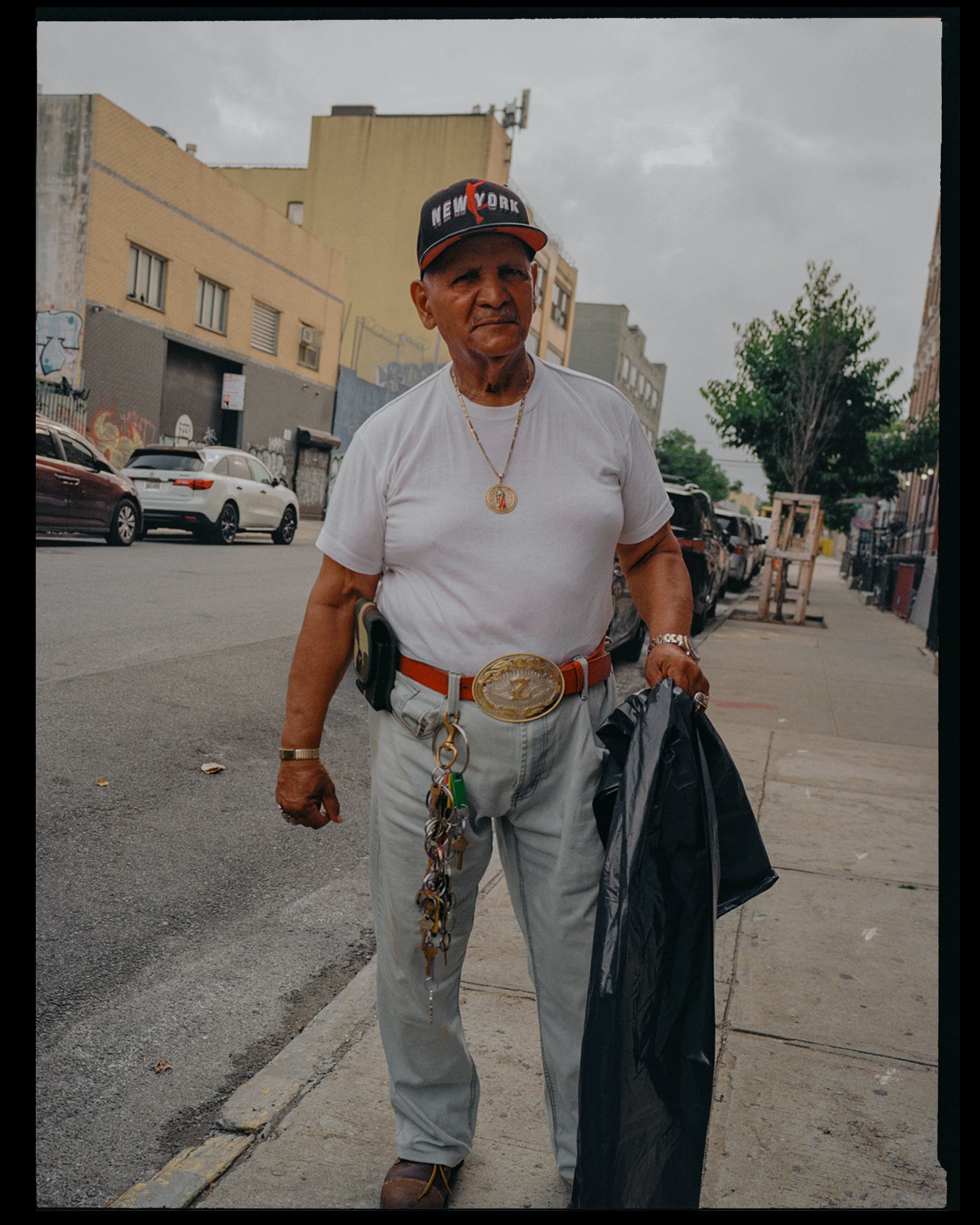 An elderly man with a serious expression wearing a black baseball cap with red and white lettering, a white t-shirt, a gold chain, and holding a black trash bag on a city sidewalk with parked cars and multistory buildings in the background.