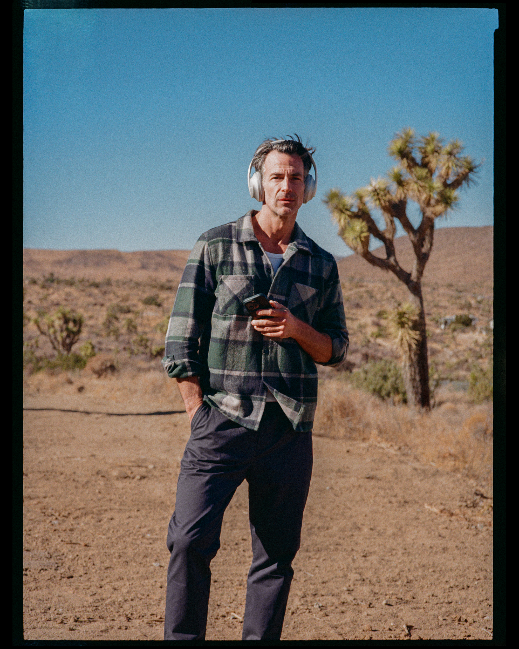 A man in a plaid shirt and dark pants wearing headphones and holding a cellphone, standing outdoors in a desert landscape with a clear blue sky and a desert plant in the background.