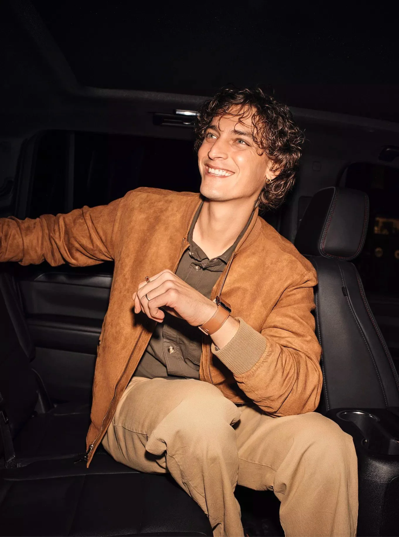 A young man with curly hair, wearing a brown suede jacket and beige pants, sitting in the backseat of a car, smiling and looking out the window.