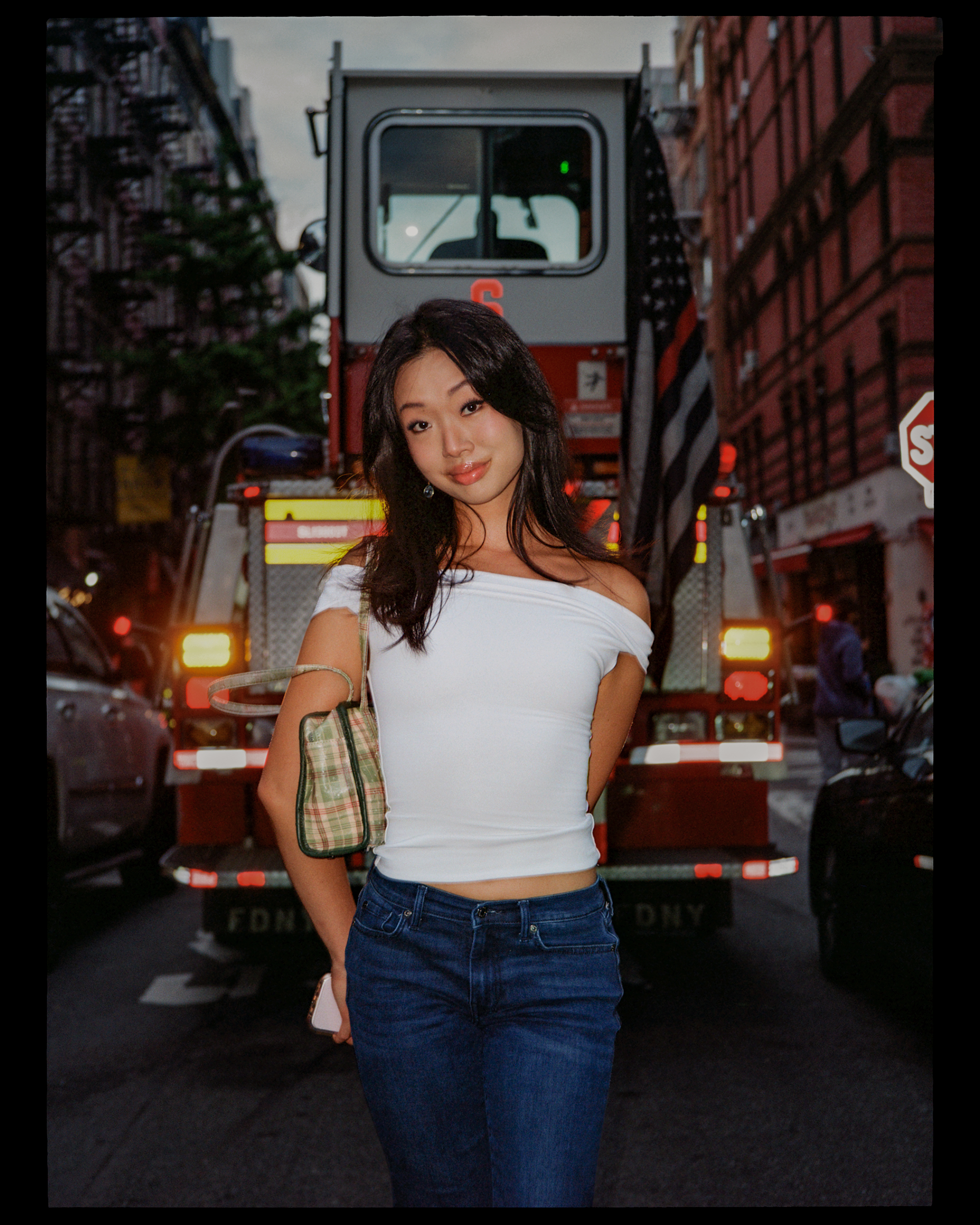 Young woman with long dark hair, wearing a white off-the-shoulder top and dark blue jeans, standing on a city street with a firetruck in the background.