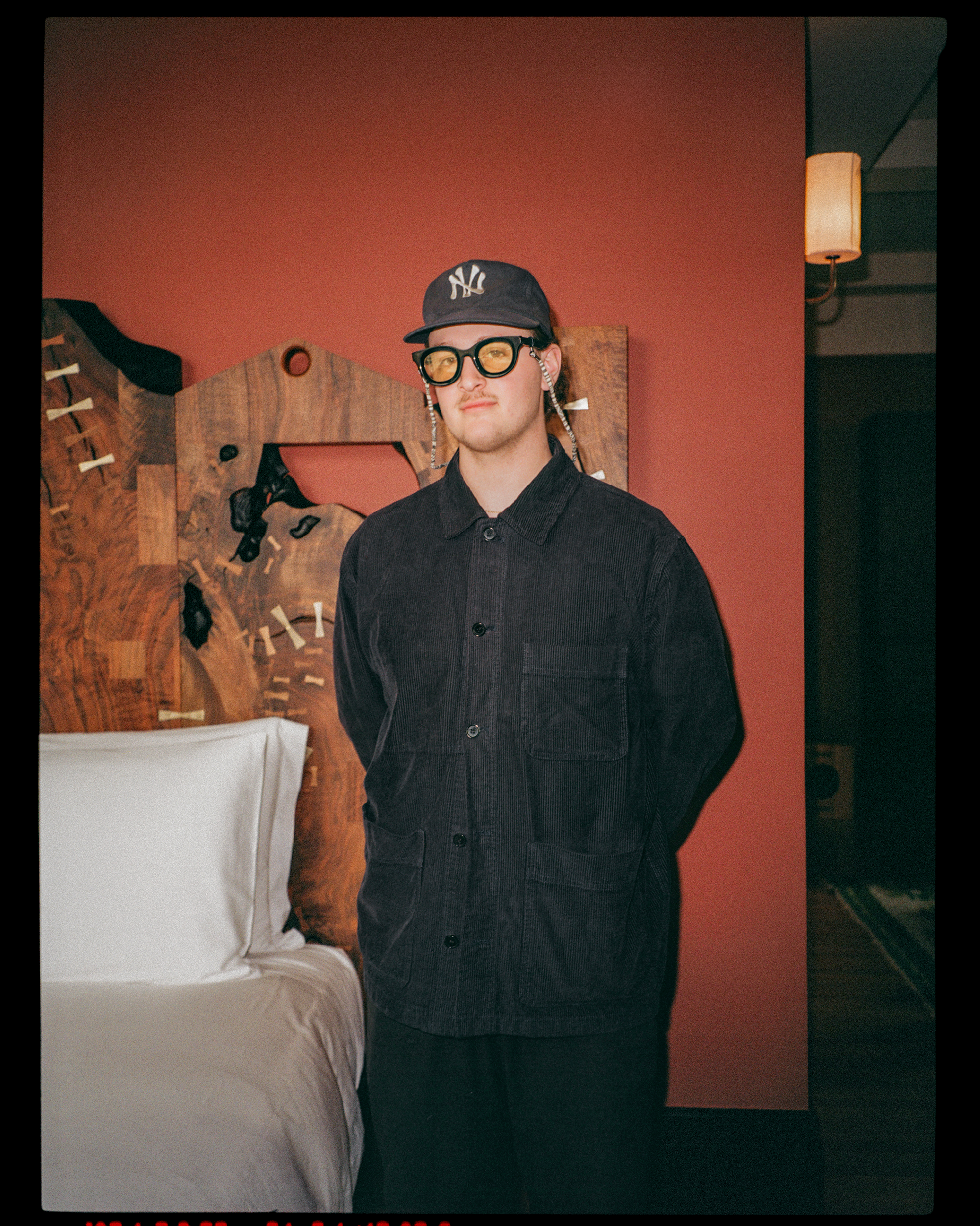 A young man with light skin, wearing a black jacket, a New York Yankees cap, and oversized glasses, standing in a room with a red wall, next to a wooden headboard and a bed with white sheets and pillow.