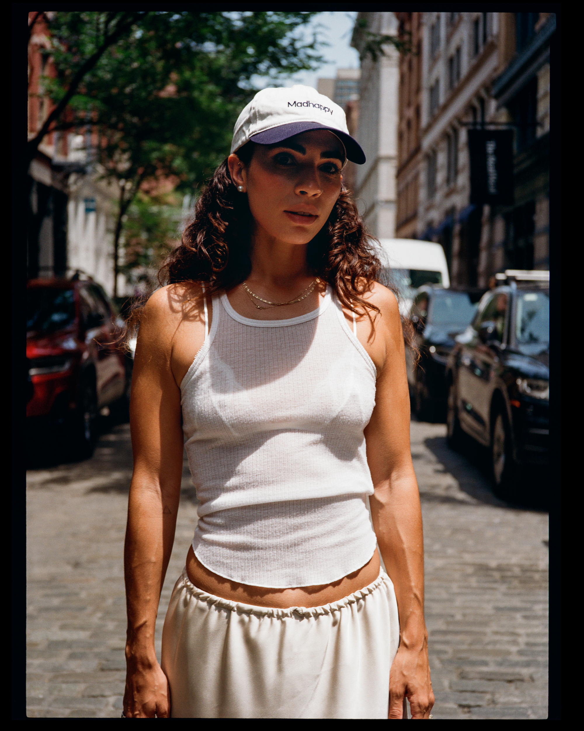 A woman with curly brown hair wearing a white tank top, a white cap with purple brim and the word 'Madhappy' on it, and pearl earrings is standing on a city street lined with parked cars and historic buildings, with trees and a partly cloudy sky in t