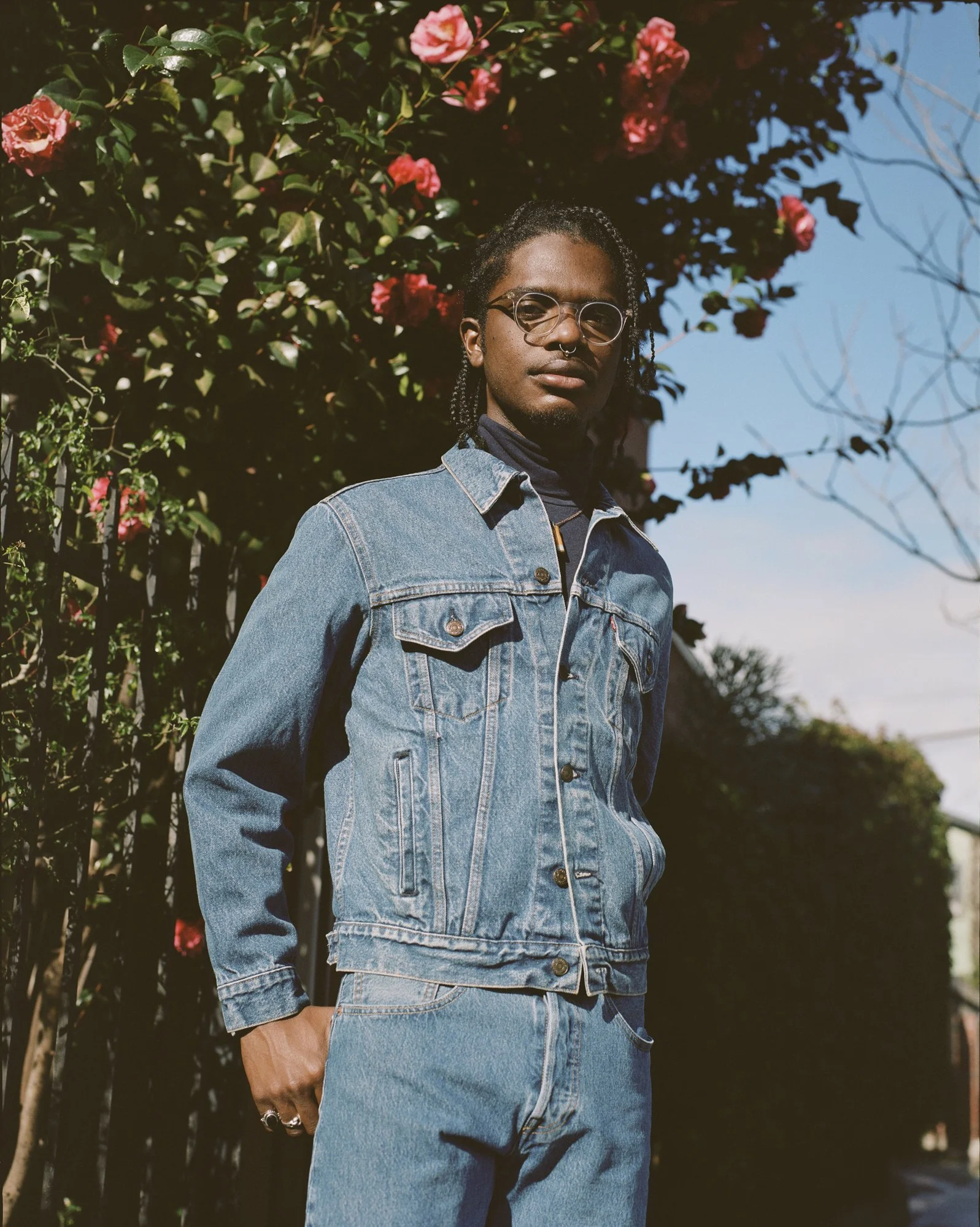 A person with dark skin, short dreadlocks, wearing glasses, a denim jacket, and jeans, standing outdoors against a backdrop of green bushes and pink flowers, with a clear blue sky.