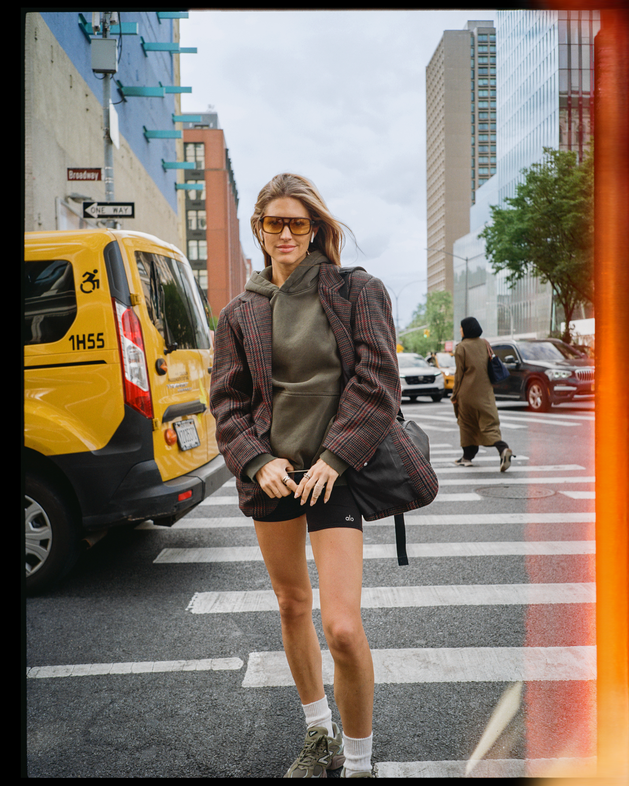 A woman with blonde hair wearing sunglasses, a hoodie, a plaid blazer, black shorts, white socks, and sneakers standing on a city street near a crosswalk.