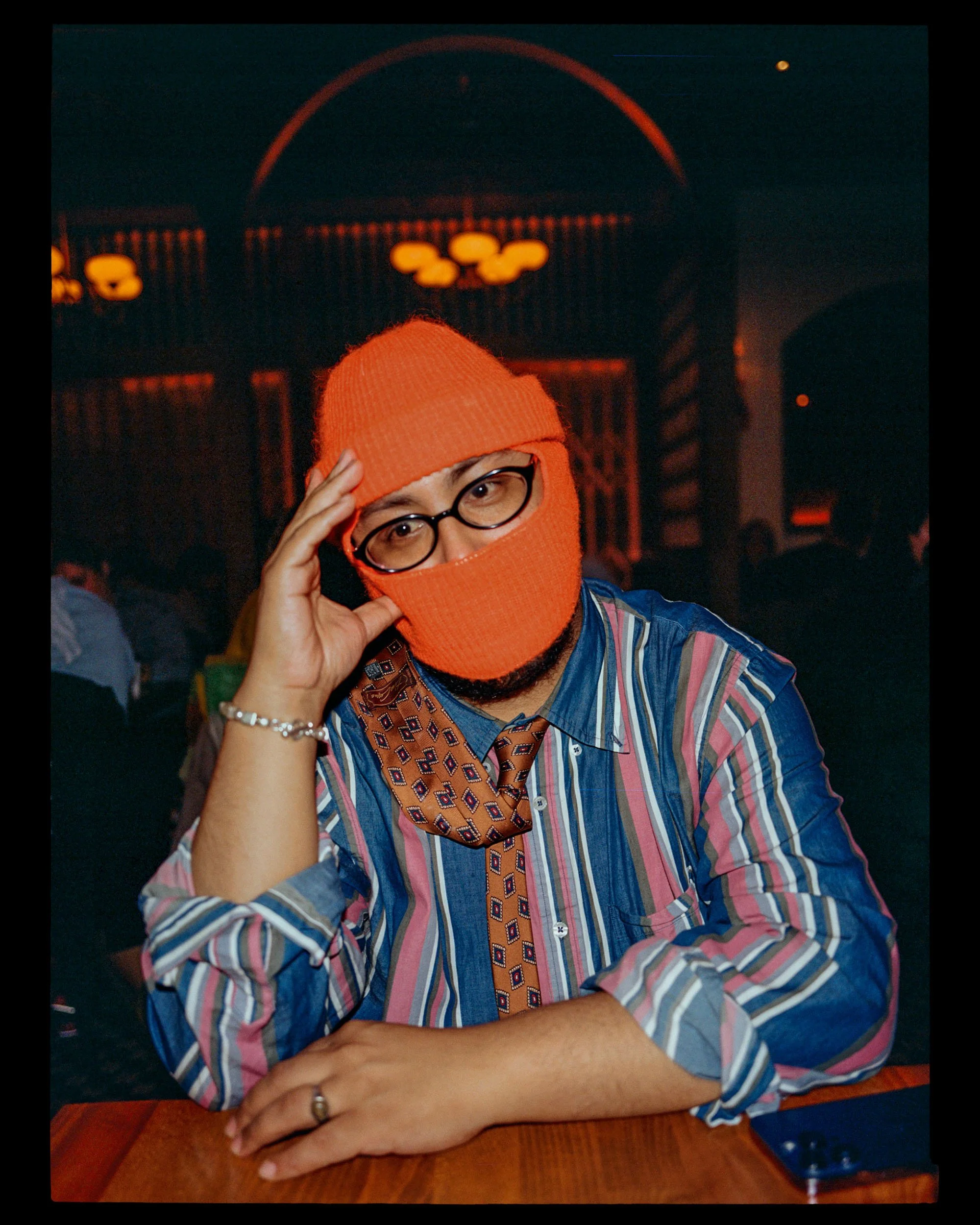 Person wearing an orange knit hat and matching face mask, glasses, and a striped shirt, sitting at a table in a dimly lit space.