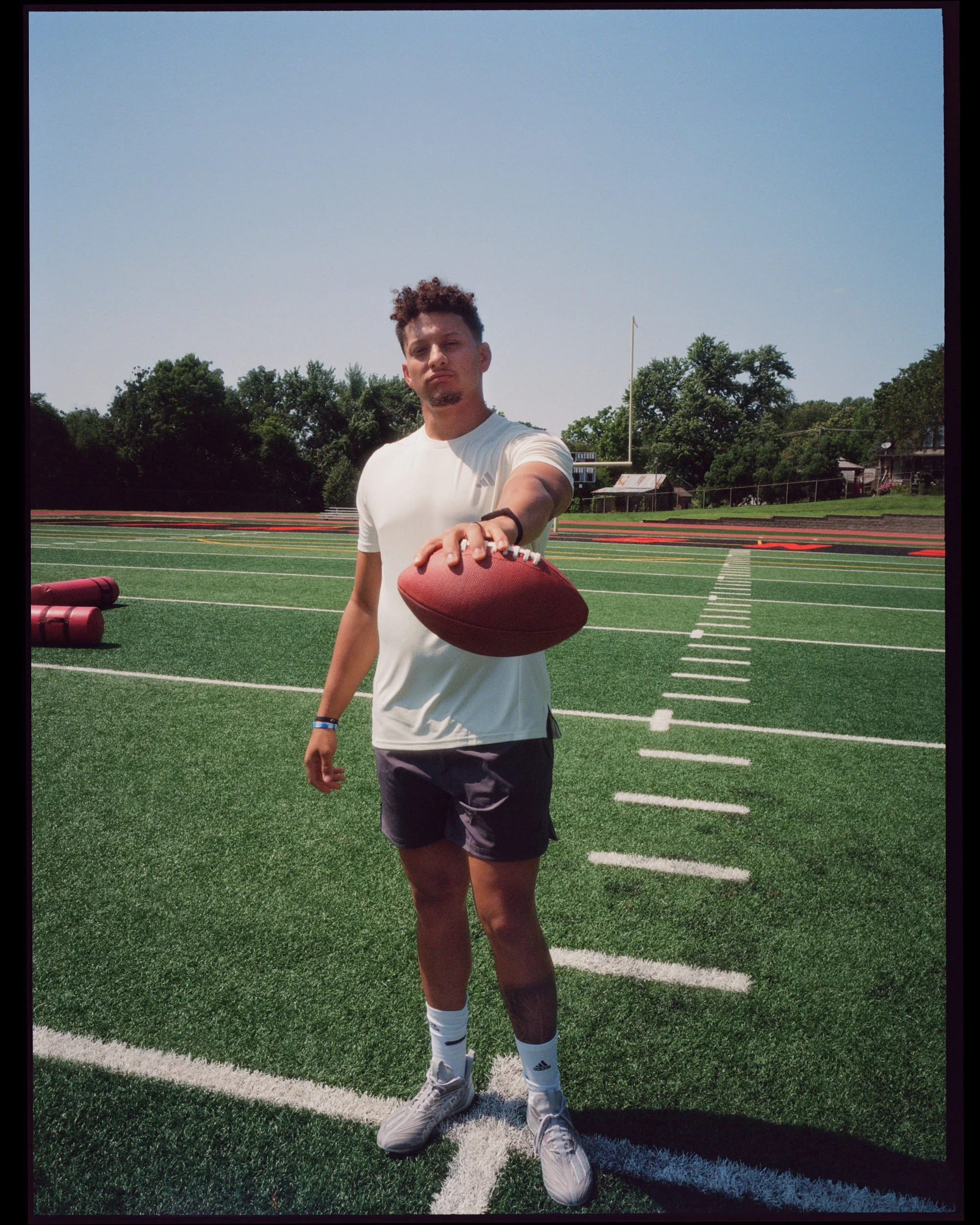 Young man stands on a football field, holding a football, wearing a white athletic shirt, black shorts, and white Nike sneakers, with a clear blue sky and trees in the background.
