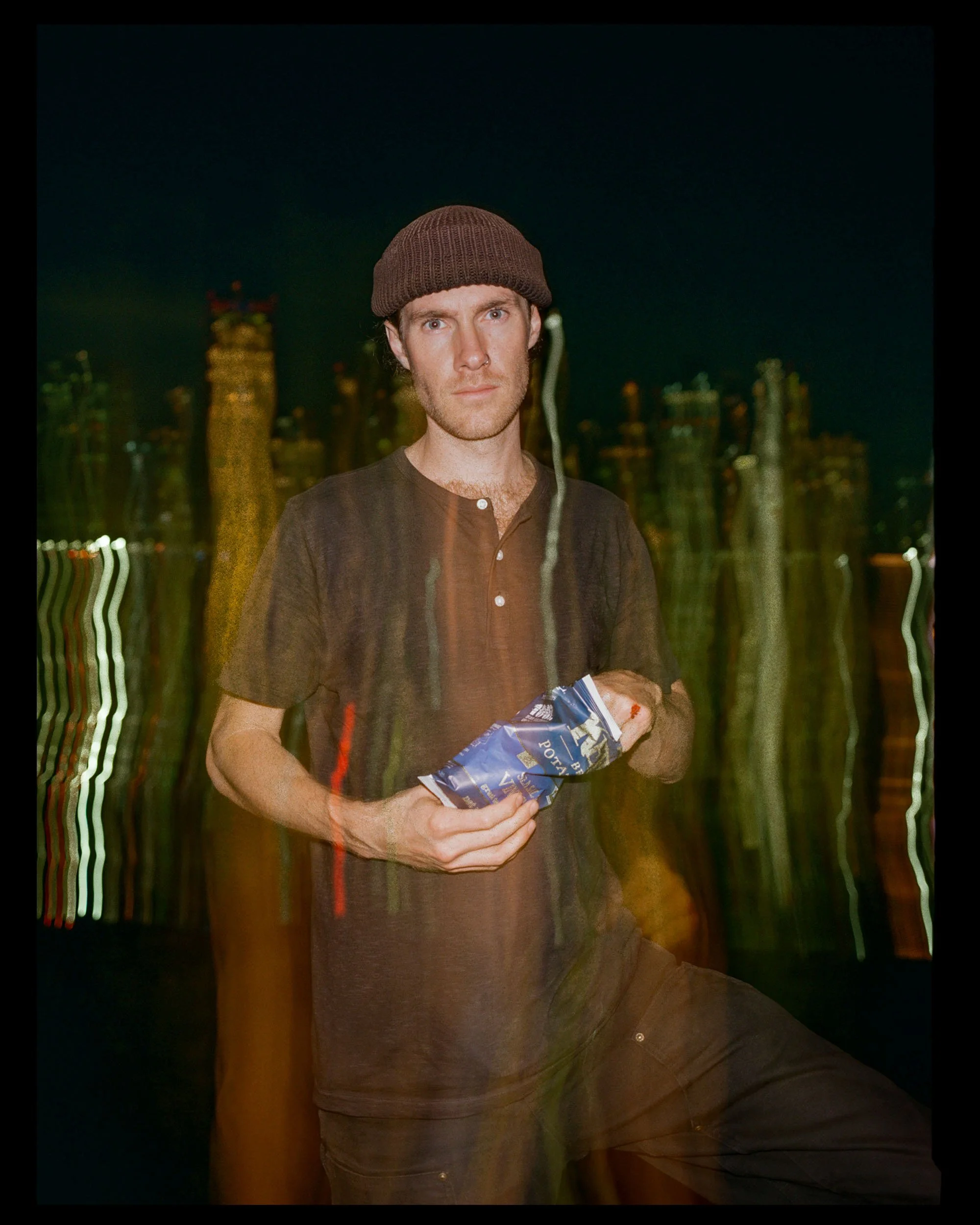 A man wearing a brown knit beanie and a dark shirt, holding a bag of Lay's potato chips, standing outdoors at night with a cityscape in the background.