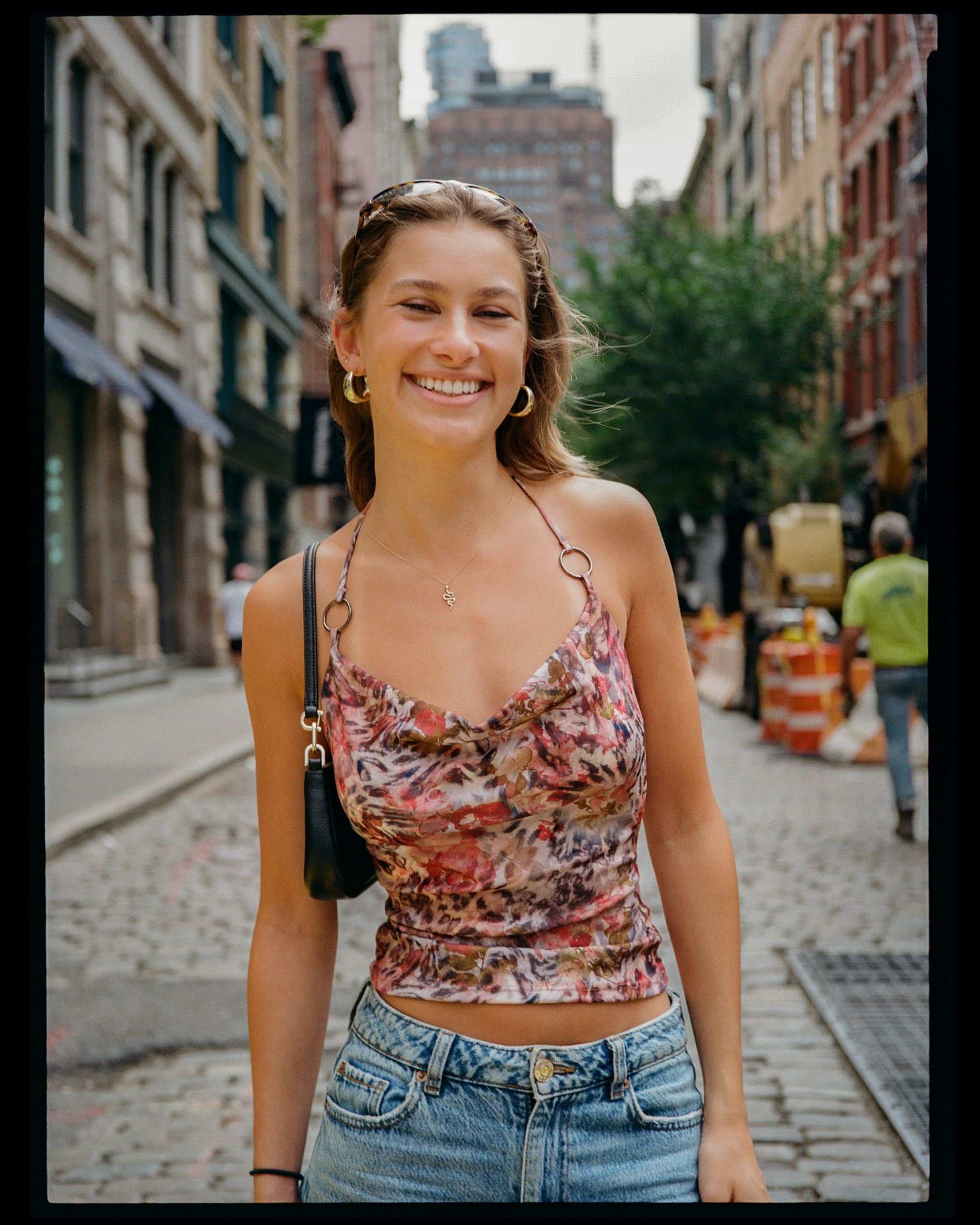 A young woman smiling outdoors on a city street with buildings and trees in the background.