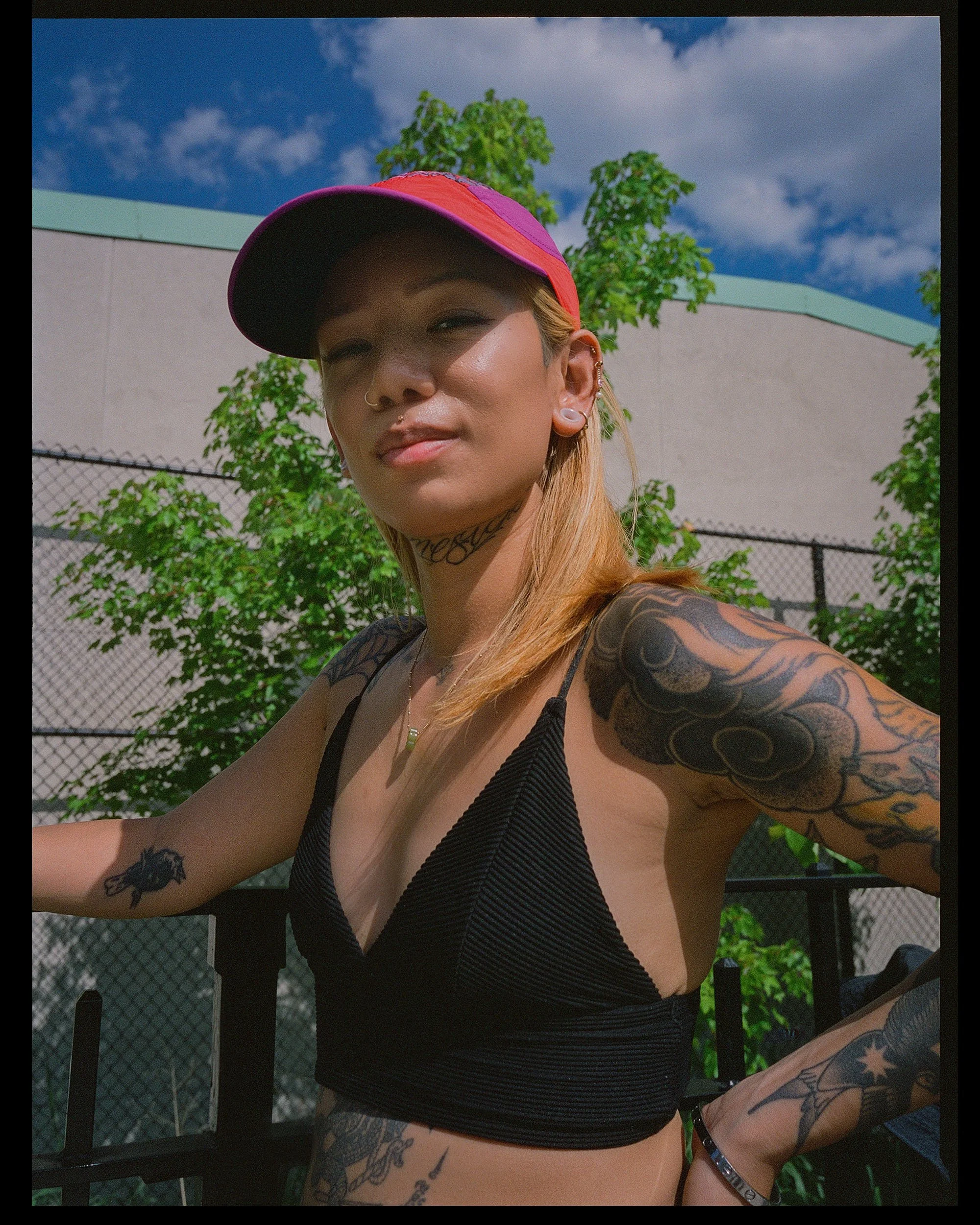 A woman wearing a red and purple cap, black crop top, and multiple tattoos, sitting outdoors on a sunny day with blue sky, clouds, green trees, and a fence in the background.
