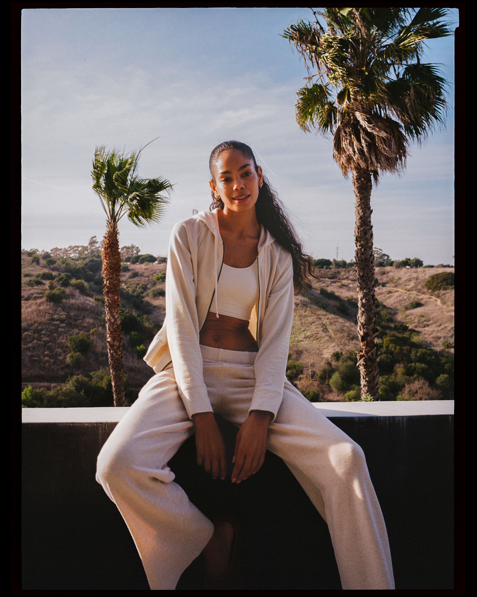 A woman sitting outdoors in front of two palm trees, wearing a beige hoodie, beige pants, and a white crop top, with a clear sky background.