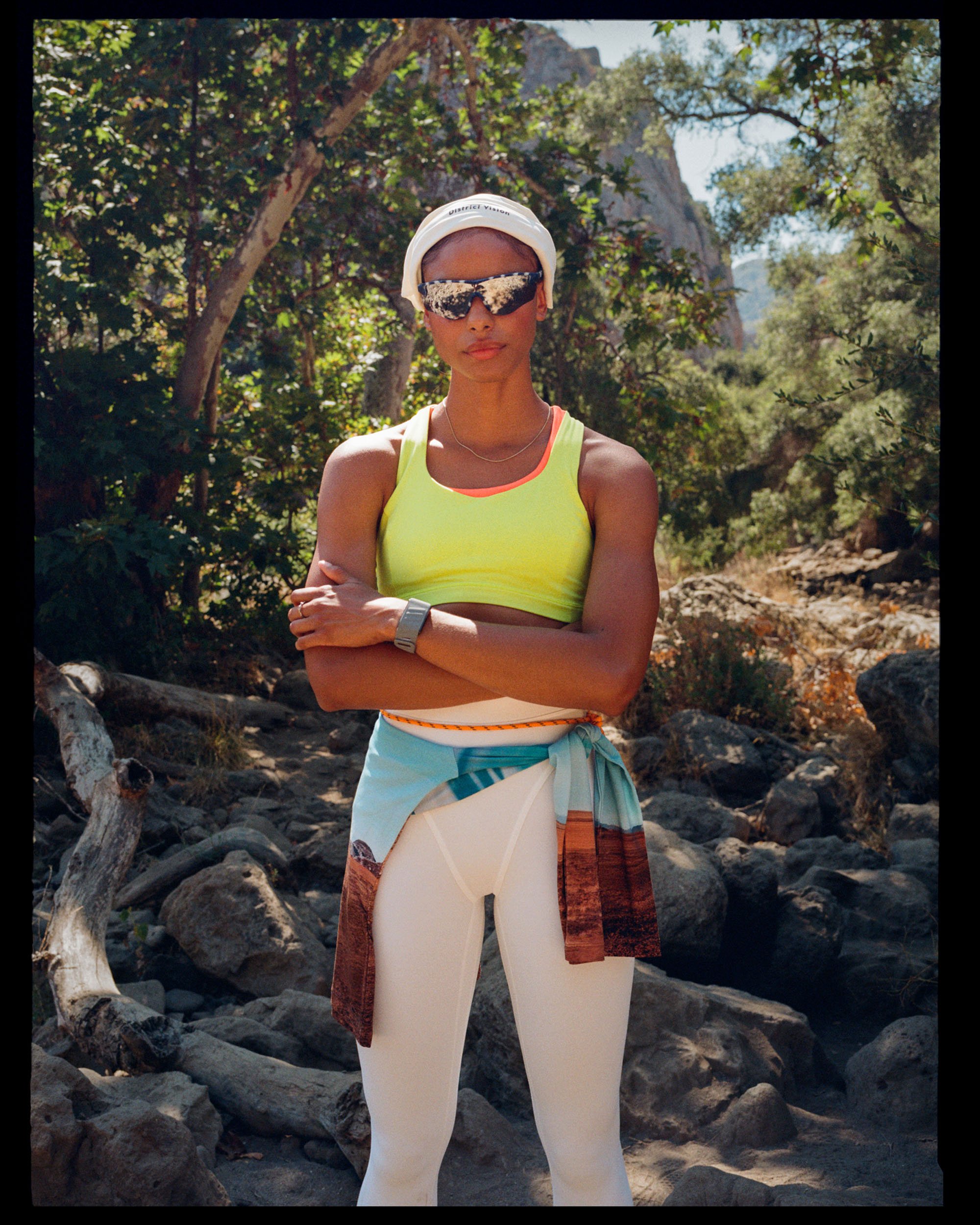 Woman standing outdoors on rocky terrain surrounded by trees, wearing a yellow athletic top, beige pants, a white hat, reflective sunglasses, a necklace, and a wristwatch.