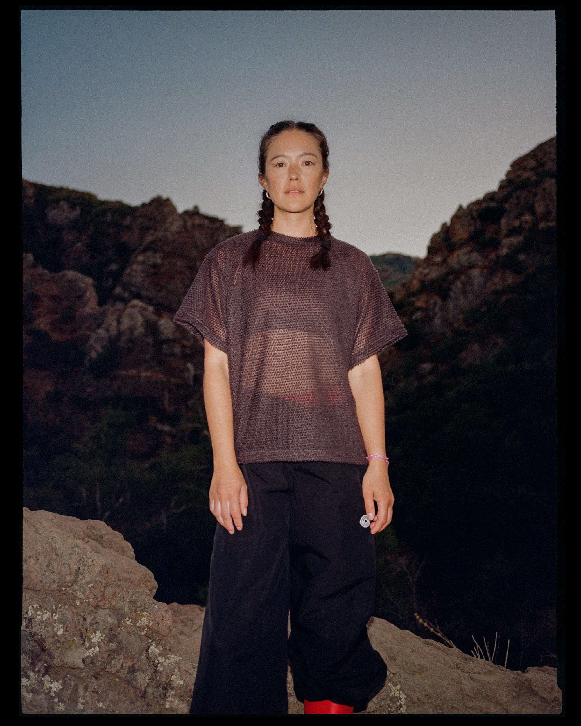A woman with braided hair standing on rocky terrain at dusk or night, wearing a loose, semi-transparent top and black pants with red accents, with large rocky formations in the background and a clear sky overhead.
