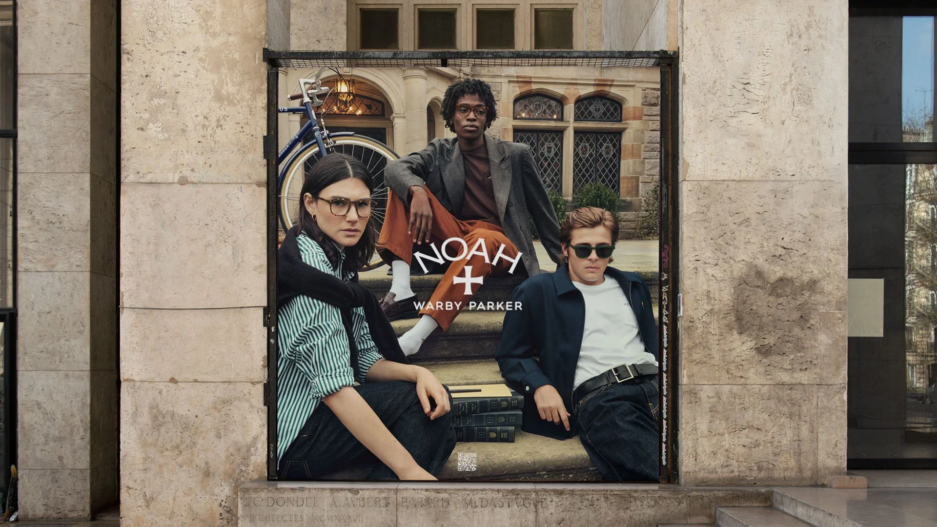 A poster on a building wall featuring three young adults posing. The person in the middle is sitting on stairs, wearing a gray blazer and orange pants. The person on the left has long dark hair, glasses, and is wearing a black and white striped shirt. The person on the right has short brown hair, sunglasses, and is wearing a navy jacket and white t-shirt. The poster includes the text "NOAH" and "WARBY PARKER".