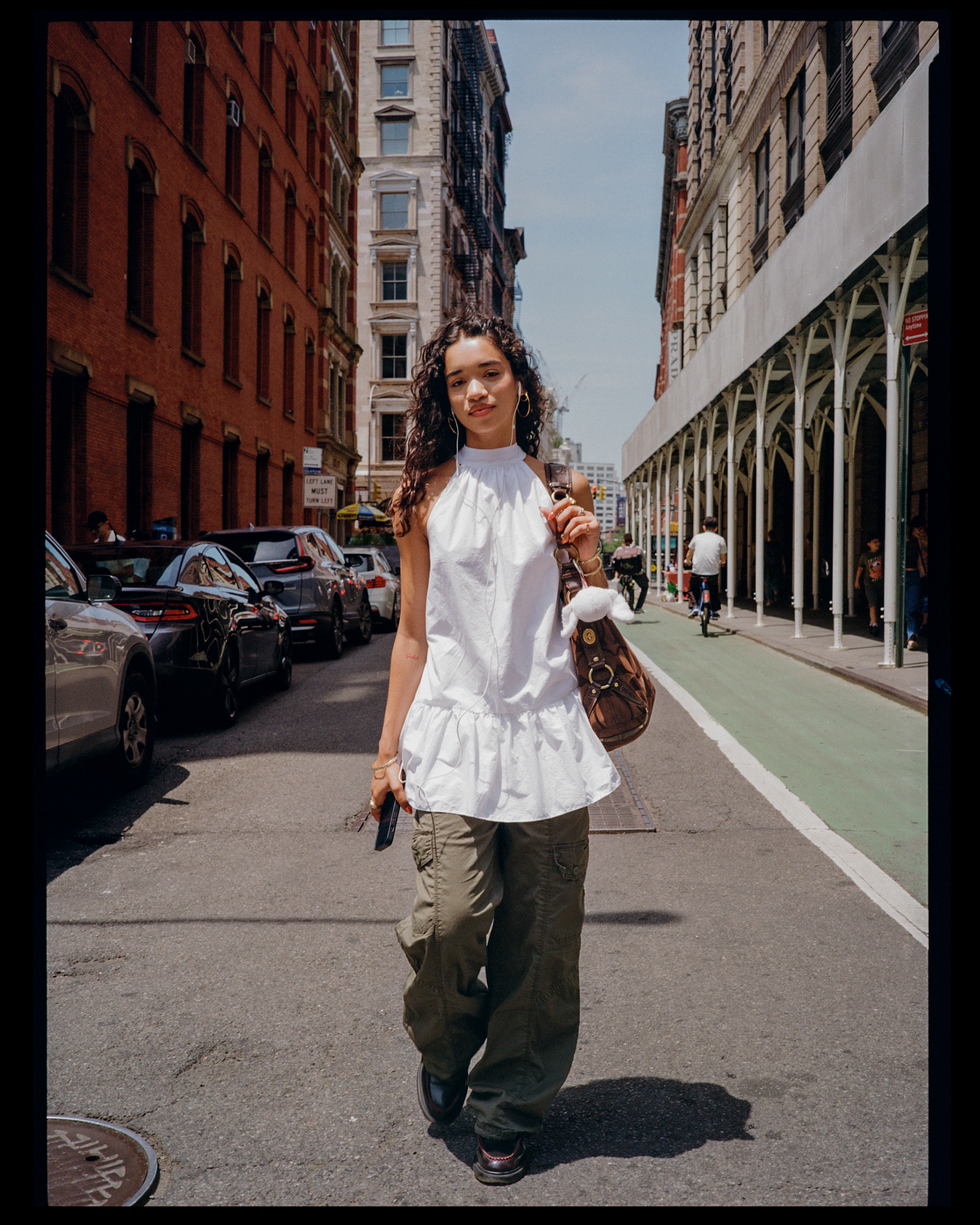 A young woman with curly dark hair walking on a city street, wearing a white sleeveless top, cargo pants, carrying a brown shoulder bag, and listening to earphones. Buildings and parked cars line the street, with a few pedestrians in the background.