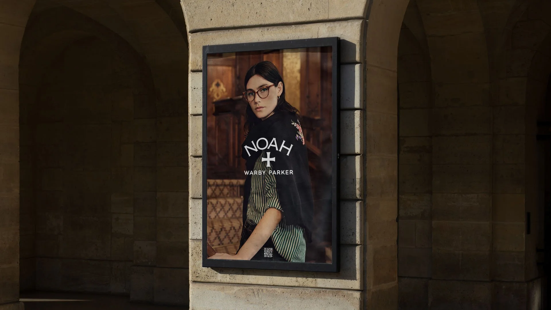 Poster of a woman with glasses and dark hair in a wooden room. The poster has the words 'NOAH' with a cross underneath and 'WARBY PARKER' at the bottom.