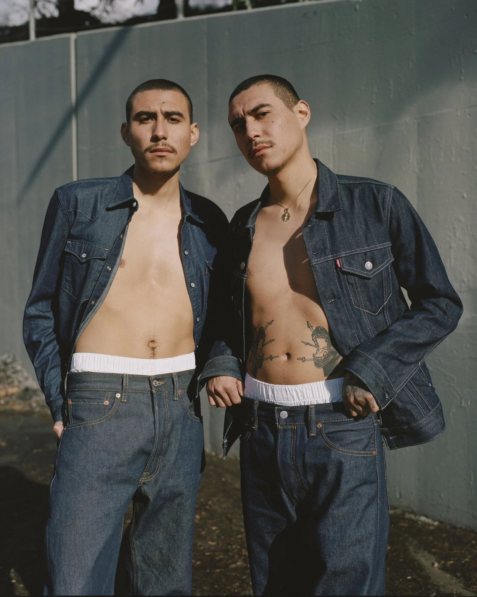 Two young men pose shirtless with their open denim jackets, standing against a gray concrete wall outdoors during the daytime. They have short dark hair and serious expressions.