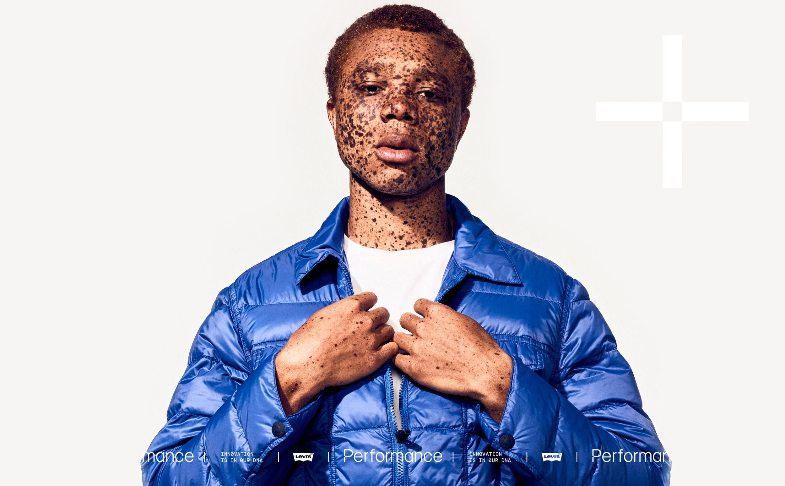 A person with freckled skin and short hair wearing a blue jacket and white shirt, holding the jacket's collar, posed against a plain background.