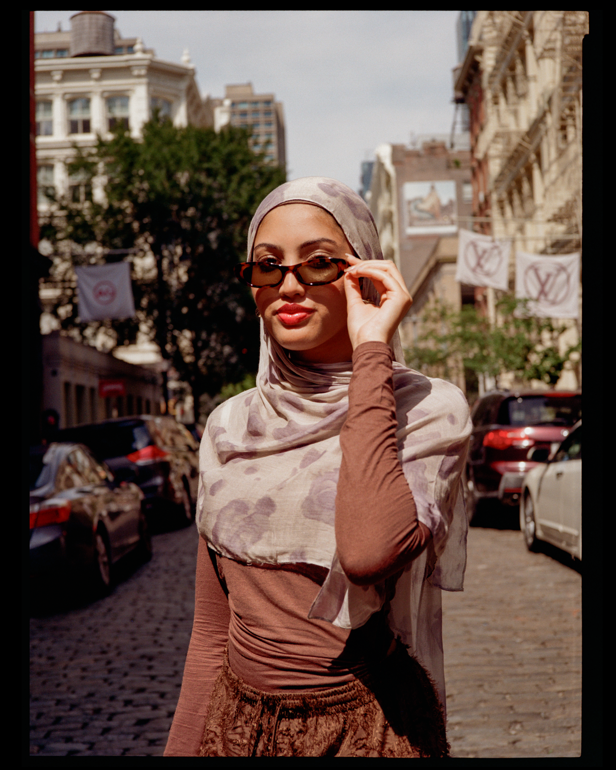 A woman wearing a patterned headscarf and sunglasses, holding the sunglasses with one hand, standing on a city street with buildings and parked cars in the background.