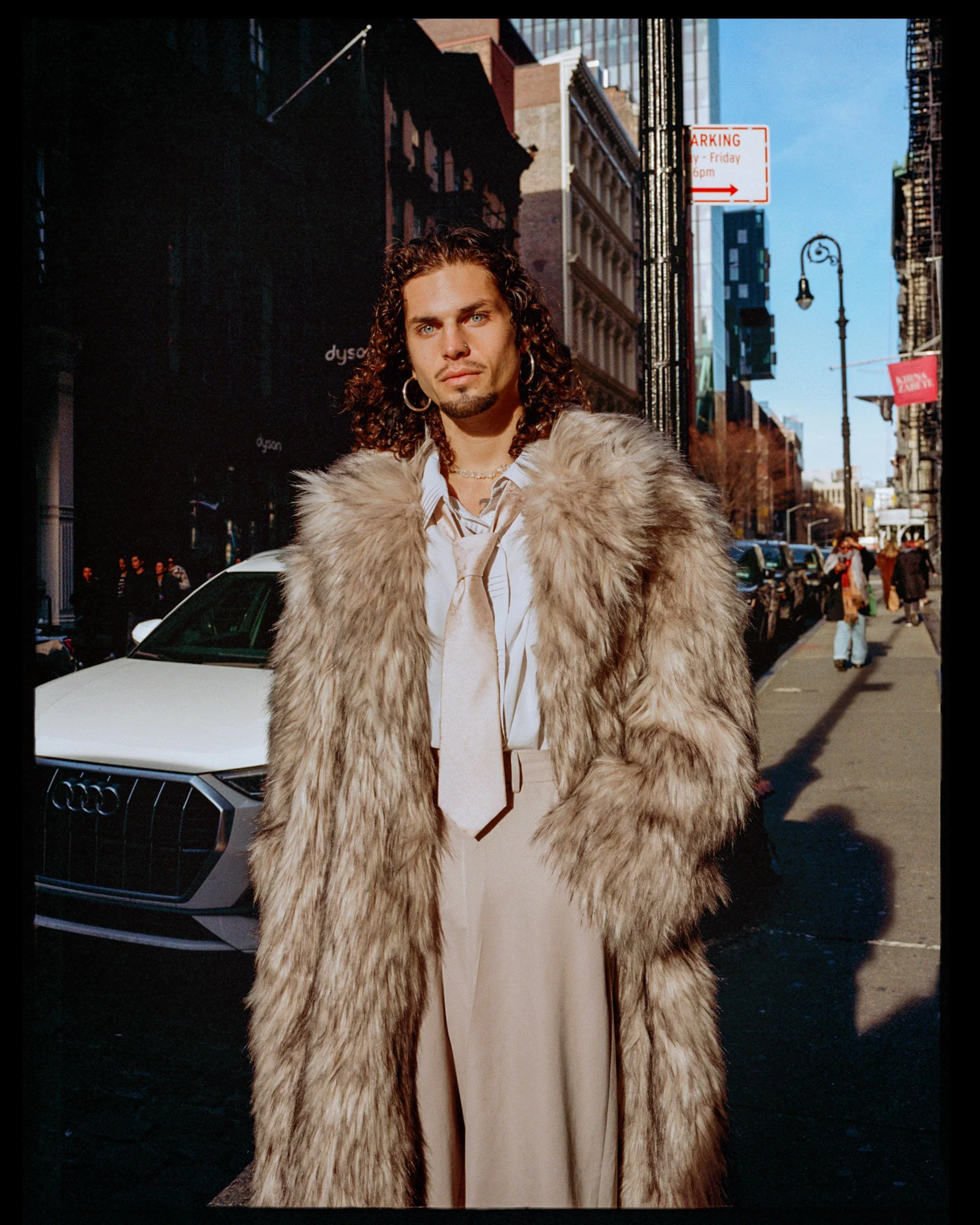 A person with long curly hair, wearing a large fur coat, earrings, and a tan tie, standing on a city street with tall buildings, a street sign, and a clear blue sky in the background.