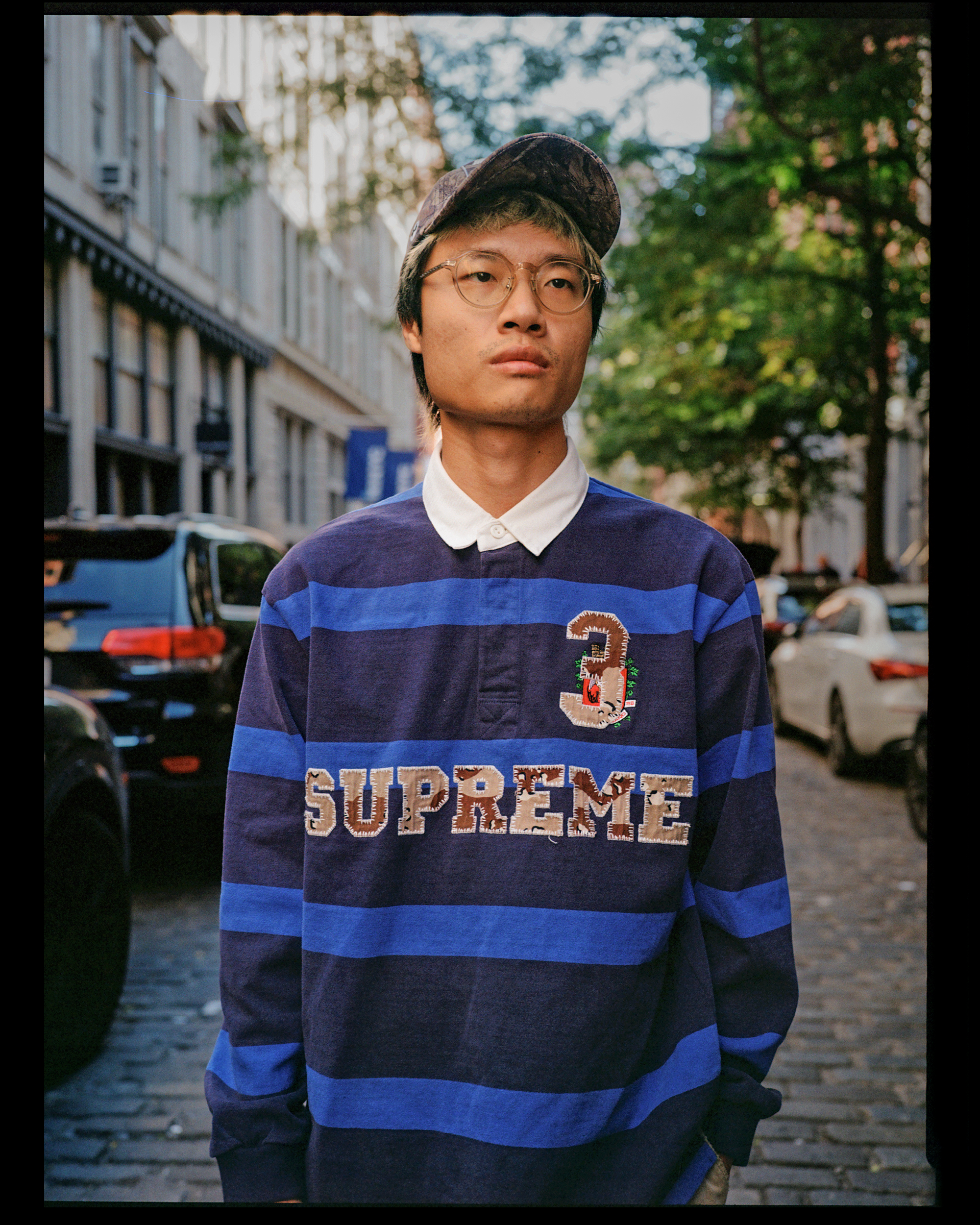 Close-up of a young man with glasses and a camo cap, wearing a navy and blue striped sweatshirt with the word 'Supreme' on it, standing outdoors on a city street with blurred cars and green trees in the background.