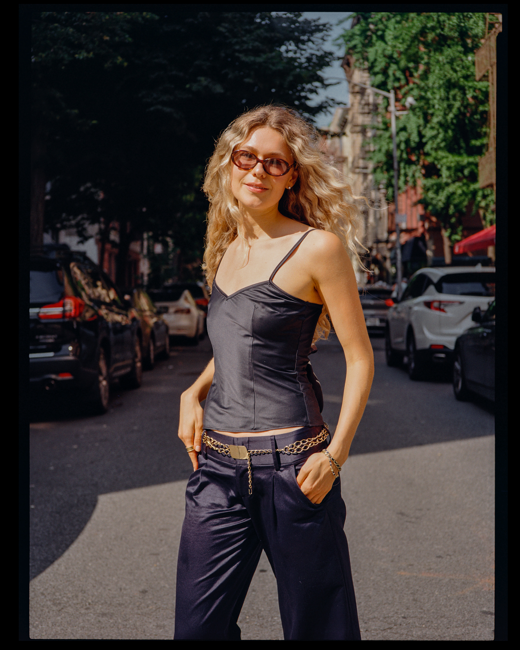 A woman with curly blonde hair wearing sunglasses, a black spaghetti strap top, and baggy black pants with a chain belt, standing outdoors on a street with cars and trees in the background.