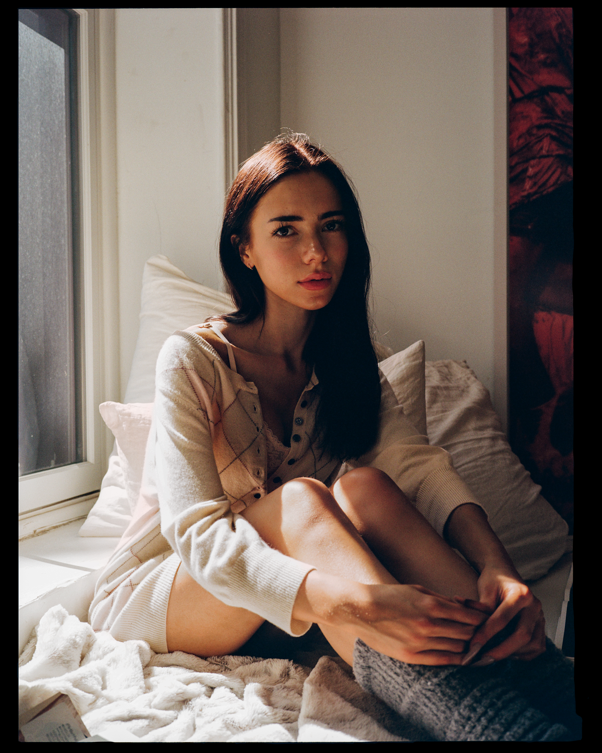 A young woman with long dark hair and light skin sitting on a bed by a window, wearing a beige sweater with a diamond pattern and shorts, with sunlight illuminating her face.