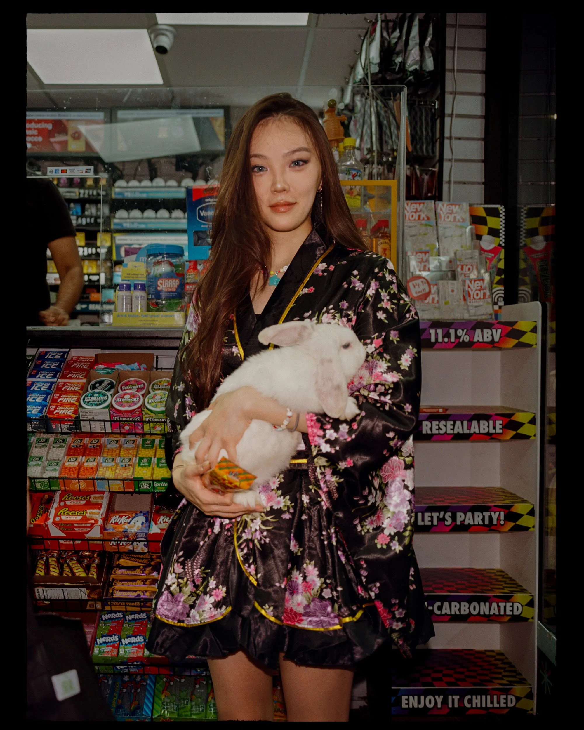 A young woman with long brown hair and fair skin wearing a black kimono with pink and purple floral patterns, holding a white bunny in her arms, standing in front of store shelves stocked with various snacks and candies.