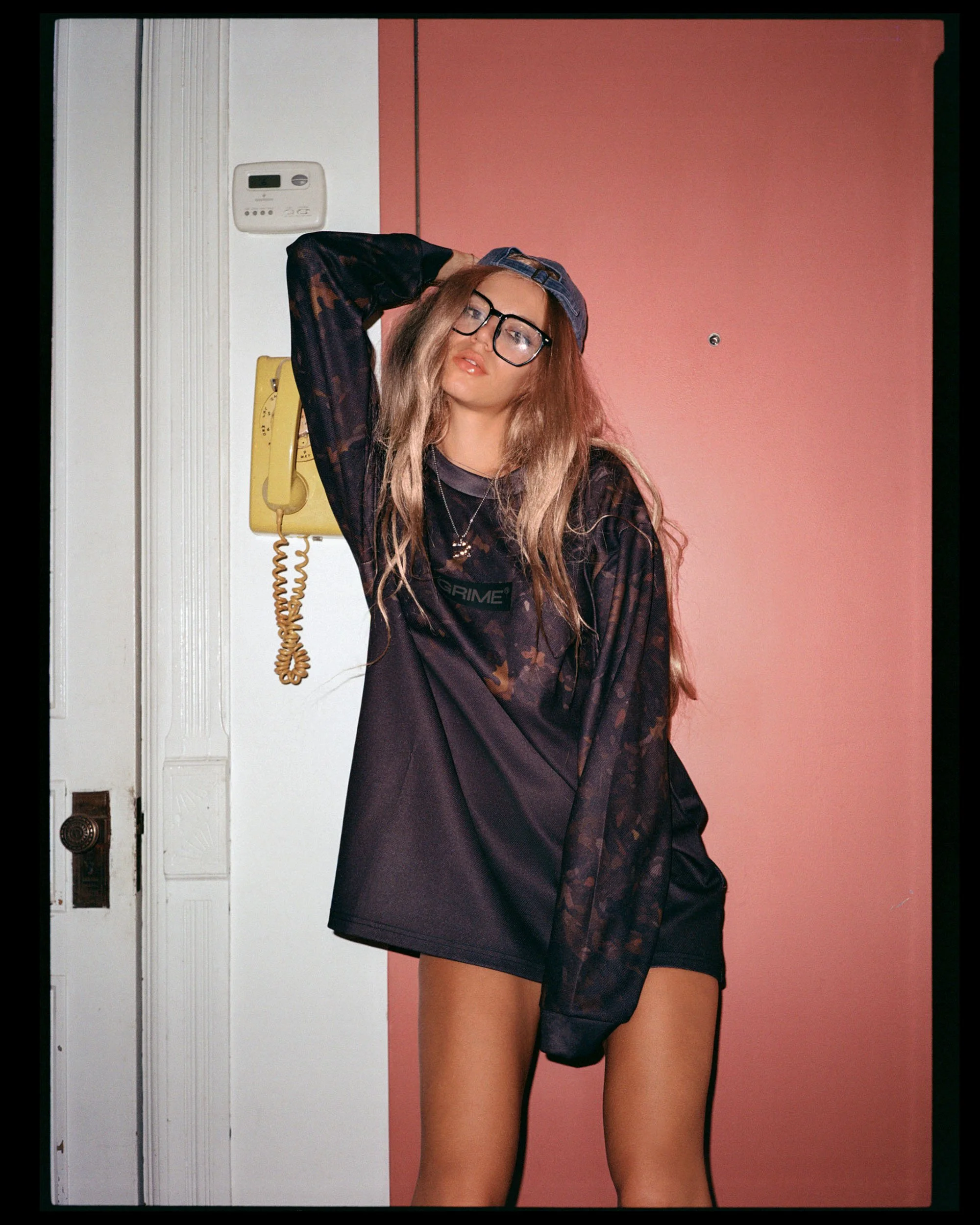 A woman with long blonde hair and glasses, wearing a blue cap and a loose black shirt, standing in front of a pink wall and a white door, with her arm raised behind her head and her face slightly turned toward the camera.