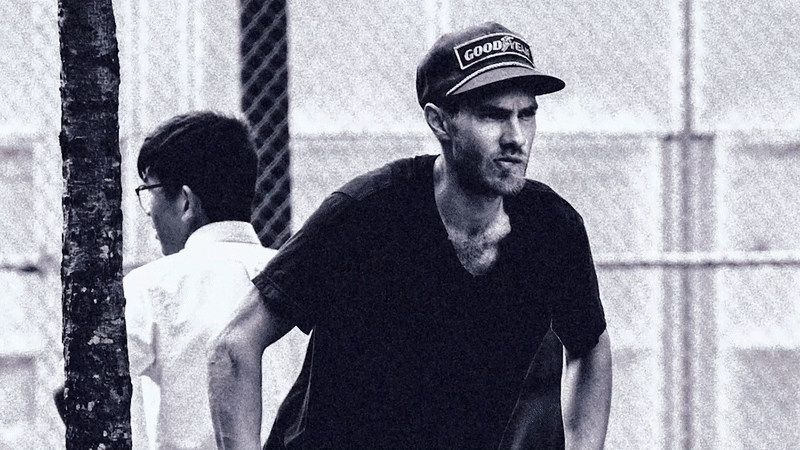 A man wearing a cap and black T-shirt walking beside a boy in glasses and a white shirt, outdoors near a textured wall and a tree.