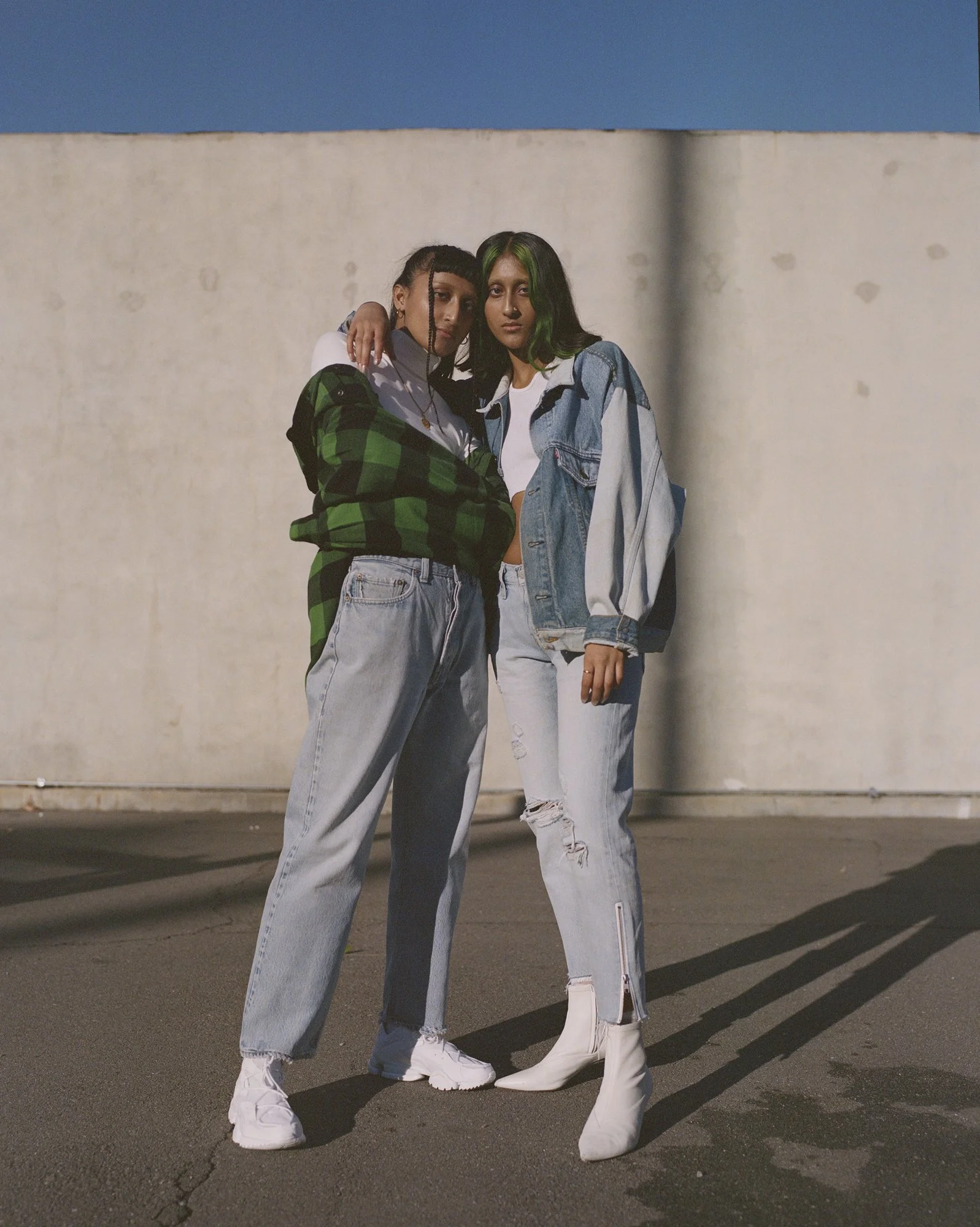 Two fashionable women standing close together in an urban parking lot at night, embracing each other and looking at camera with confident expressions. One woman has green-highlighted dark hair and wears a denim jacket and ripped jeans; the other has braided hairstyle and wears an oversized checkered shirt and baggy jeans.