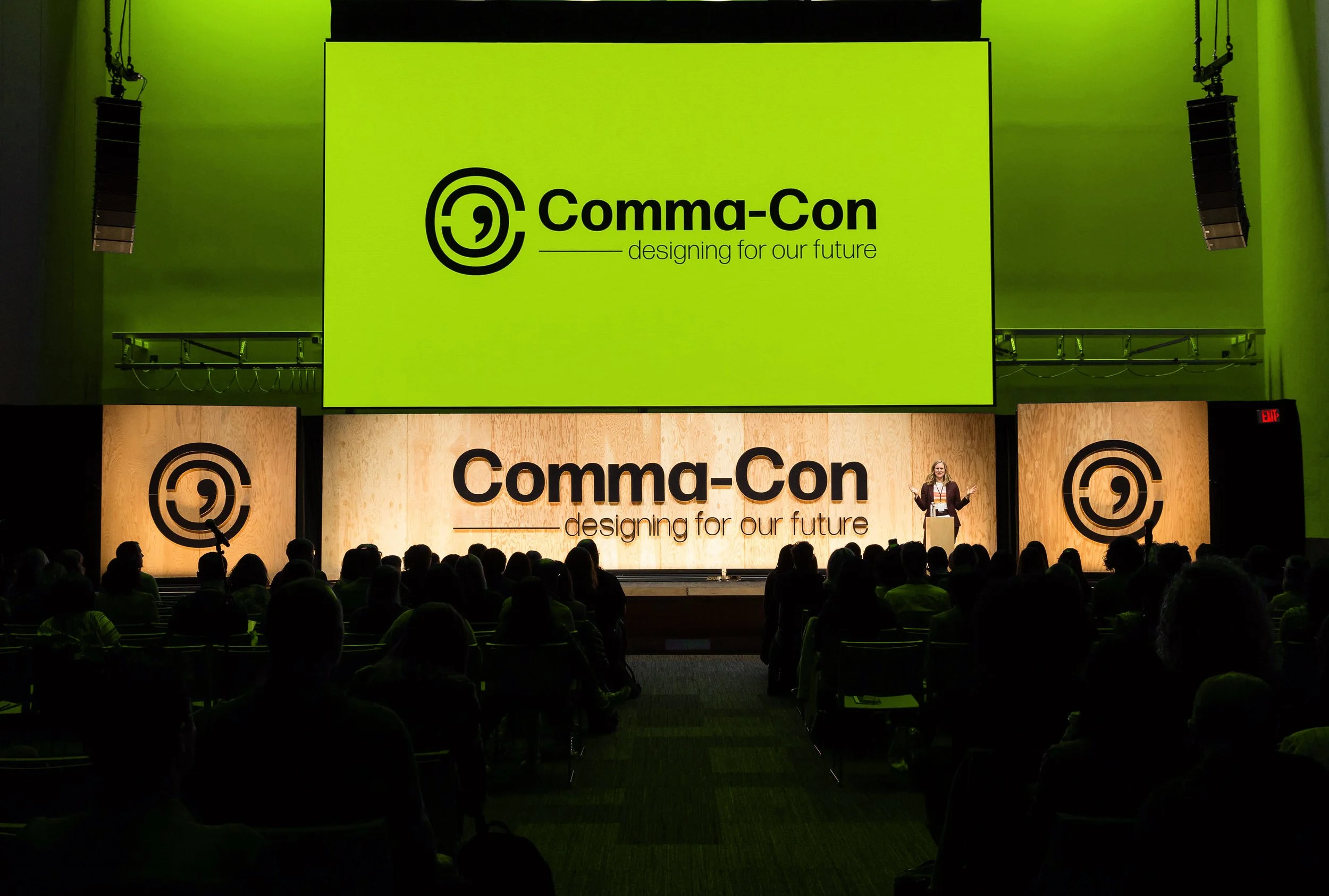 A large green screen displays the logo and slogan of Comma-Con, 'designing for our future.' A woman stands on stage in front of an audience, speaking at a conference with the same logo projected behind her, along with black speakers hanging on either side of the stage.