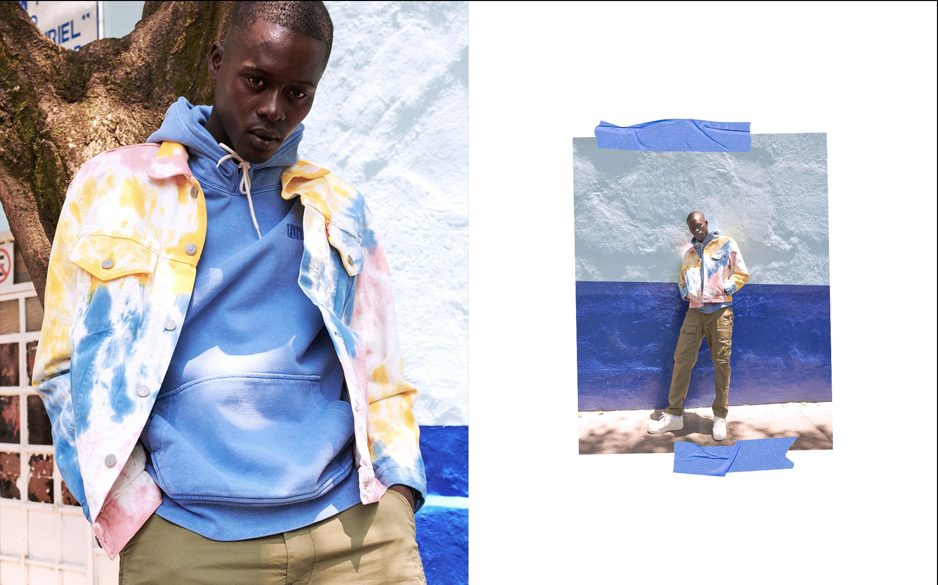 A young man wearing a colorful tie-dye jacket and blue hoodie stands outdoors in front of a painted blue and white wall, with shadows of trees on the ground.