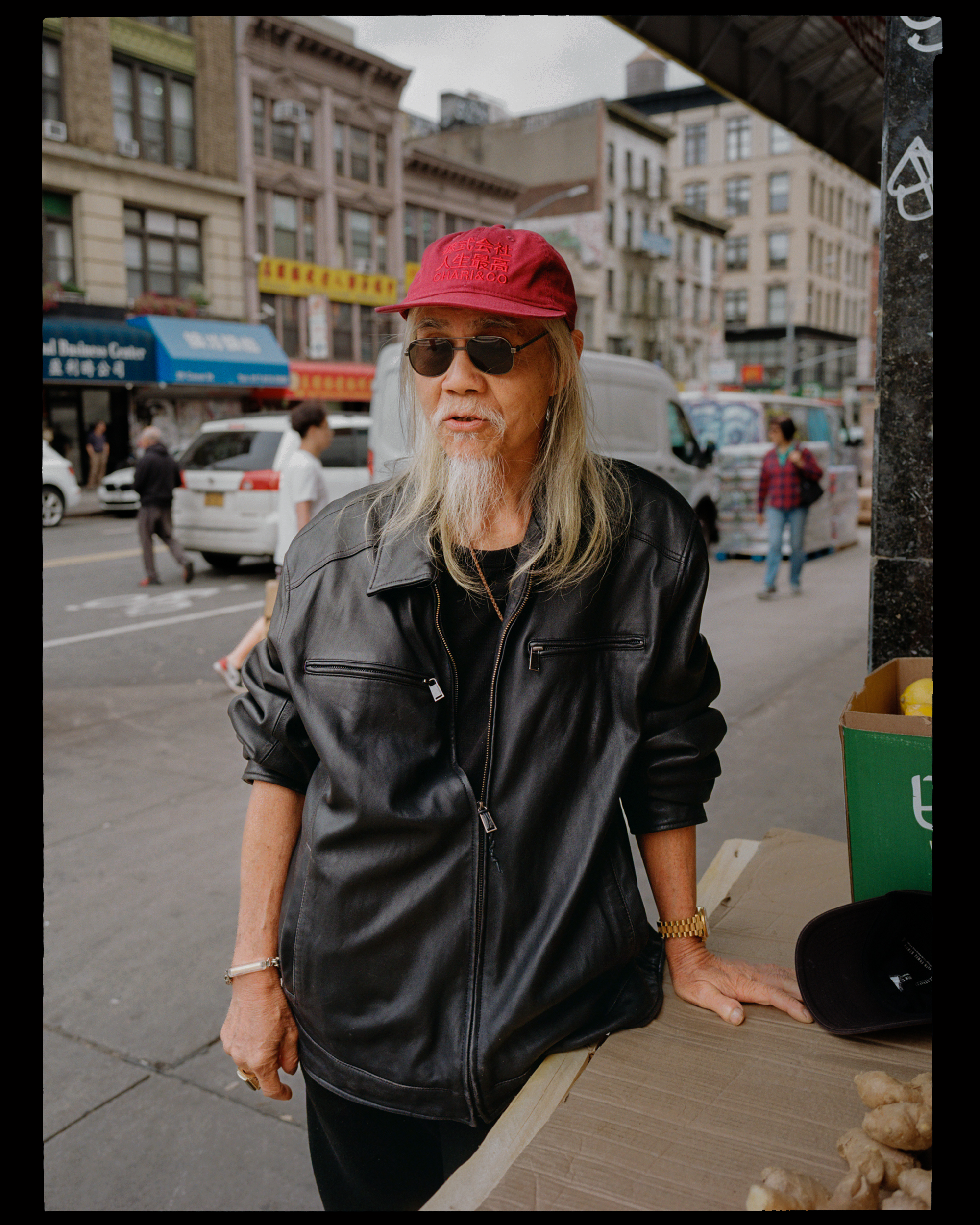 An elderly woman wearing sunglasses, a red baseball cap, and a leather jacket is standing outdoors on a city street. She is leaning on a wooden table with a green container and some garlic. The background shows pedestrians, parked cars, and buildings