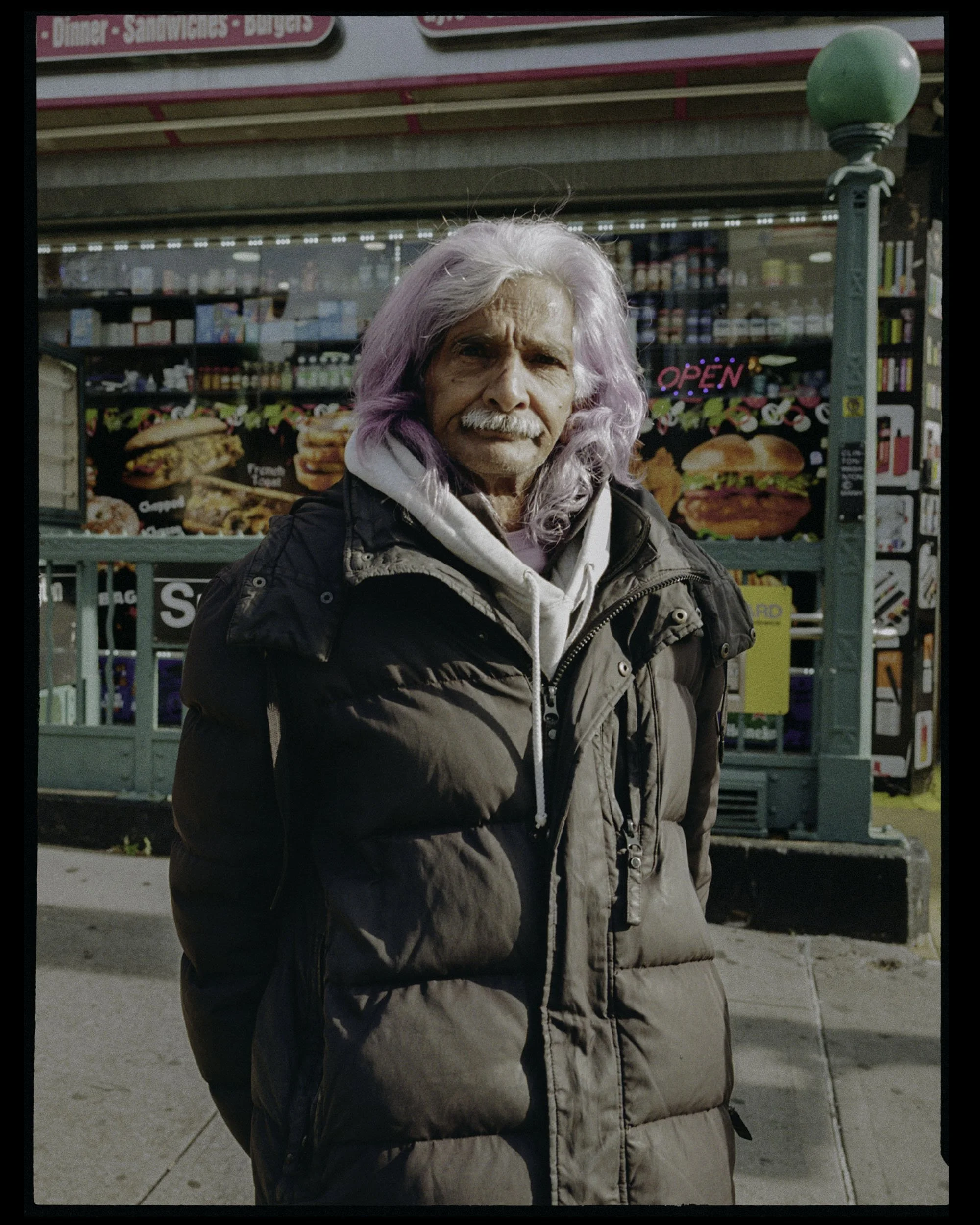 An elderly man with long, wavy, lavender-colored hair and a mustache stands outside a store with a pink and blue sign. He is wearing a tan and black jacket with a white hoodie underneath. The background shows a storefront with a neon "OPEN" sign and 