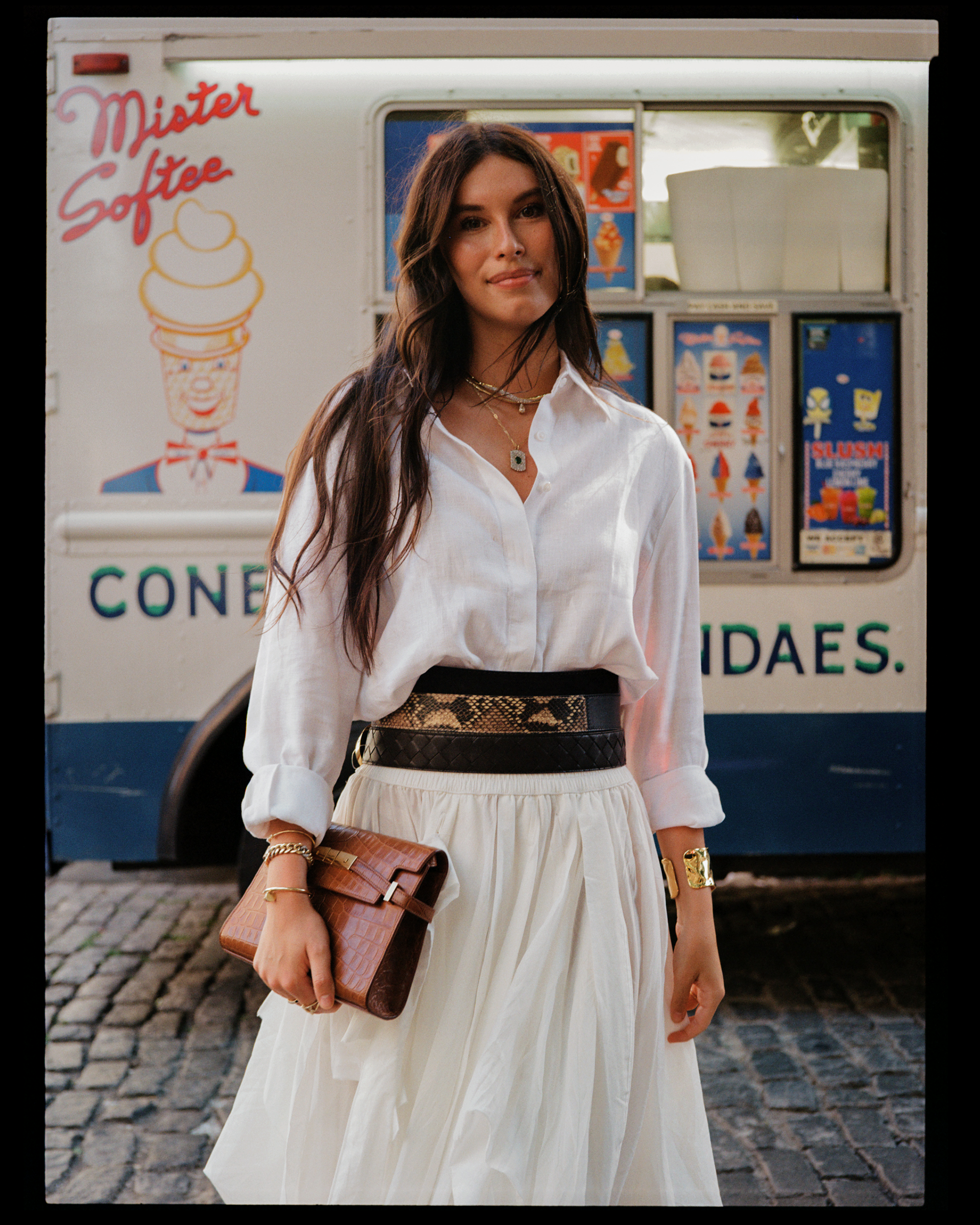 A woman standing in front of an ice cream truck, wearing a white blouse and a white skirt, holding a brown clutch purse, with jewelry including necklaces and bracelets.