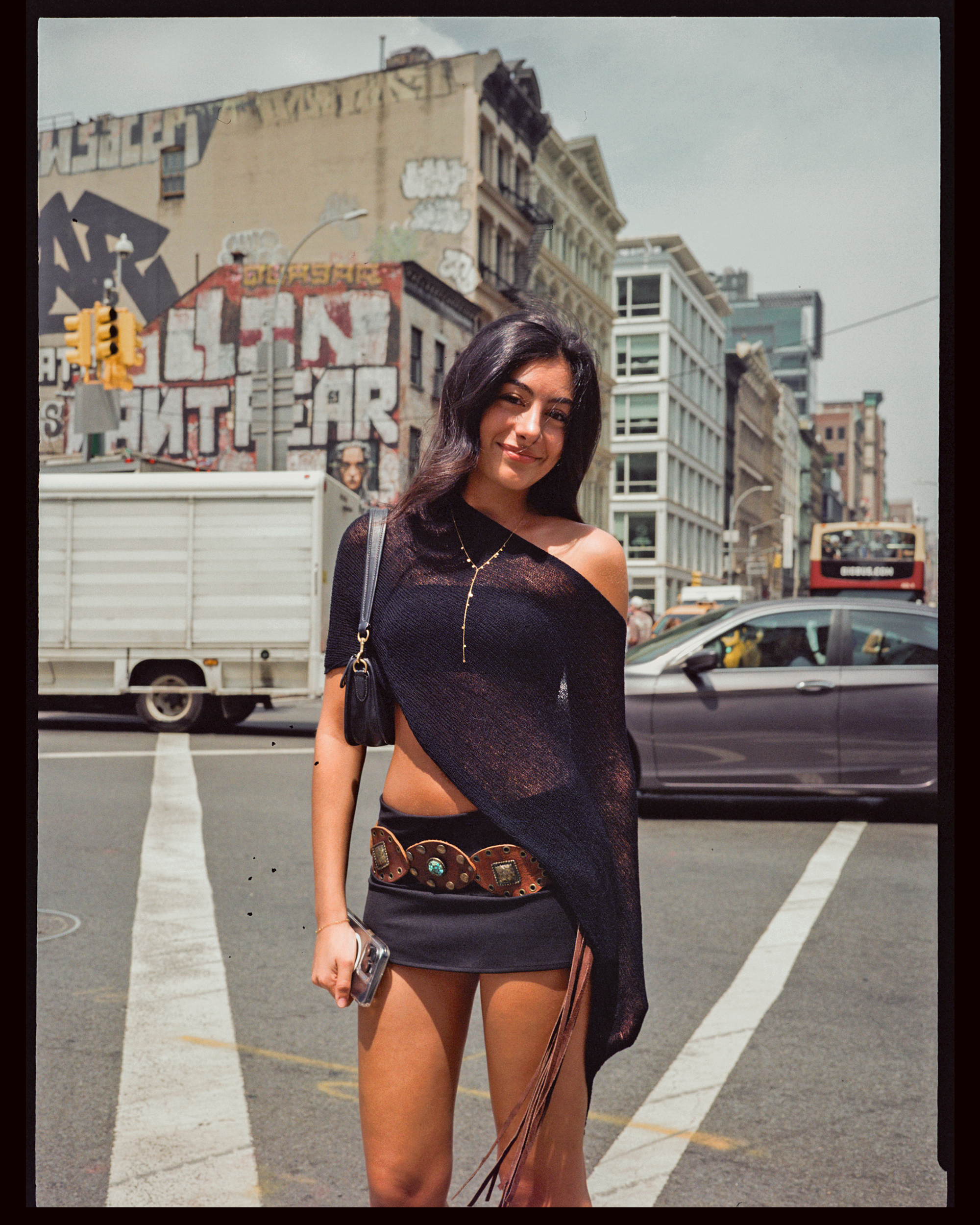 A woman with long dark hair, smiling, standing on a city street with buildings, a traffic light, and a bus in the background. She is wearing a black off-shoulder top and a dark skirt, holding a silver smartphone in her right hand, with a brown purse 