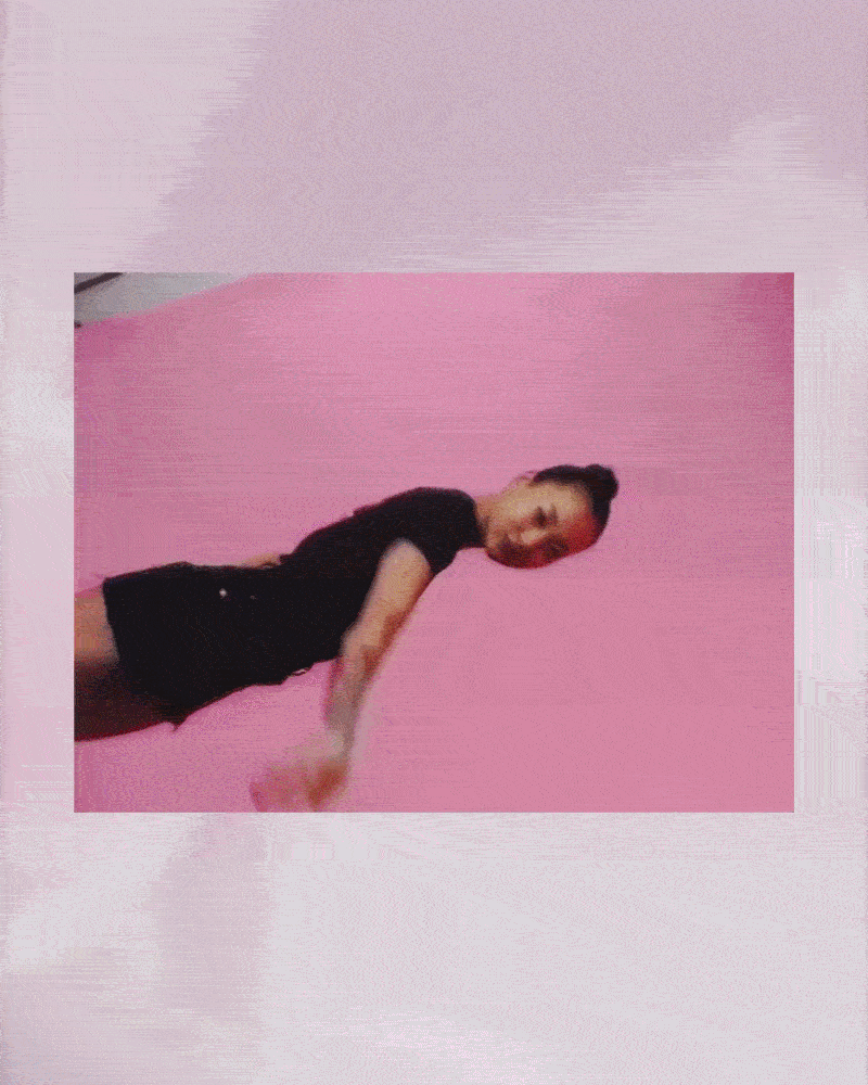 A woman with a braided bun hairstyle is performing a yoga pose against a pink wall, wearing a black shirt and black shorts.