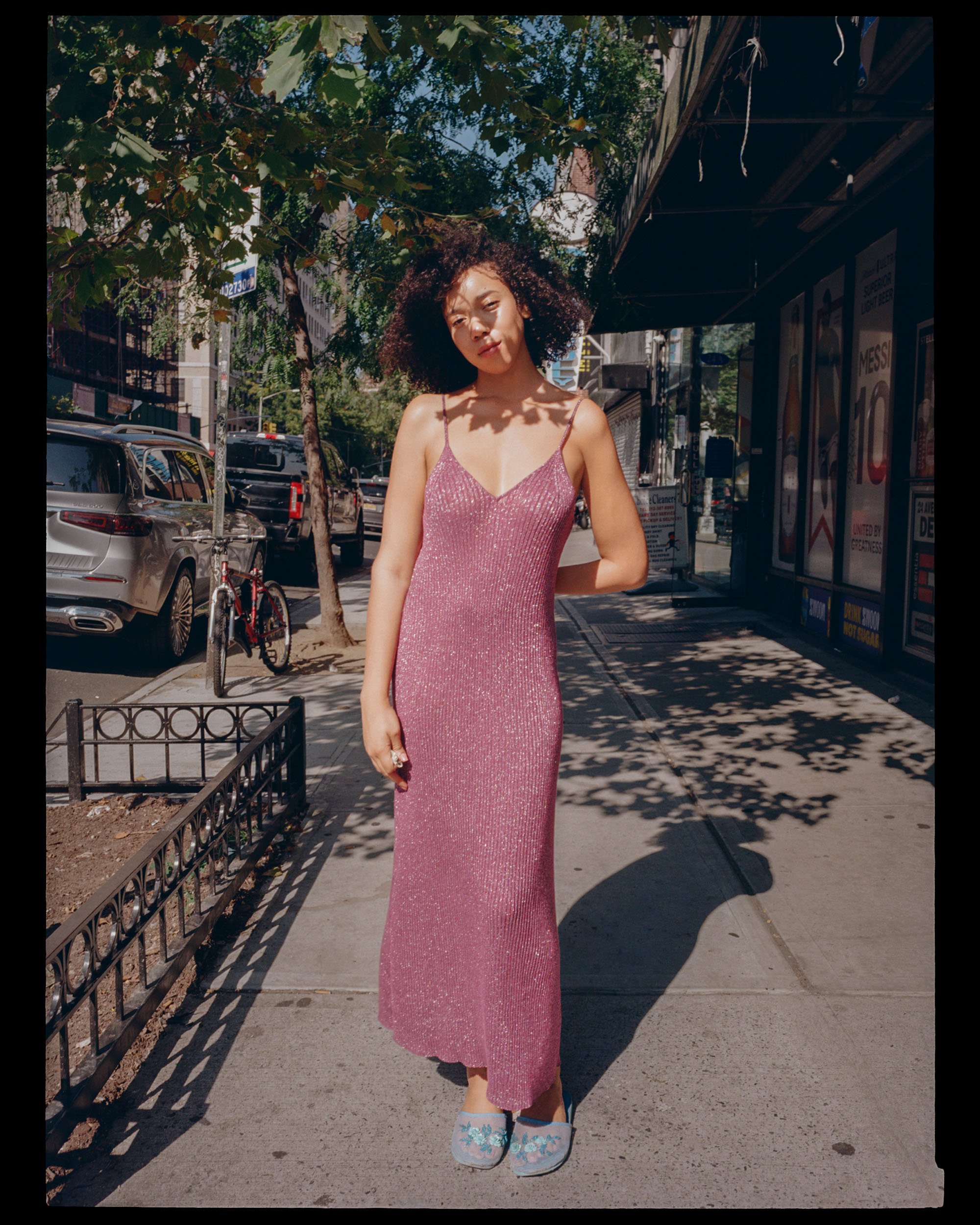 Young woman with curly hair standing on sidewalk in city street, wearing pink shimmer dress, blue floral slippers, and posing with shadow and sunlight patterns on her face and body.