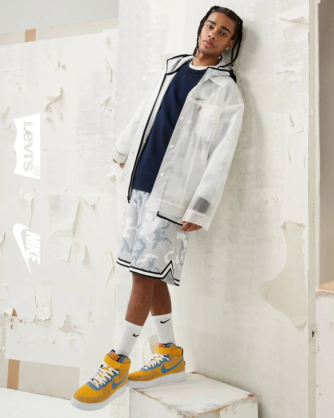 Young man with braided hair wearing a navy shirt, white transparent jacket, white camouflage shorts, white socks, and yellow Nike sneakers standing against a distressed white wall.