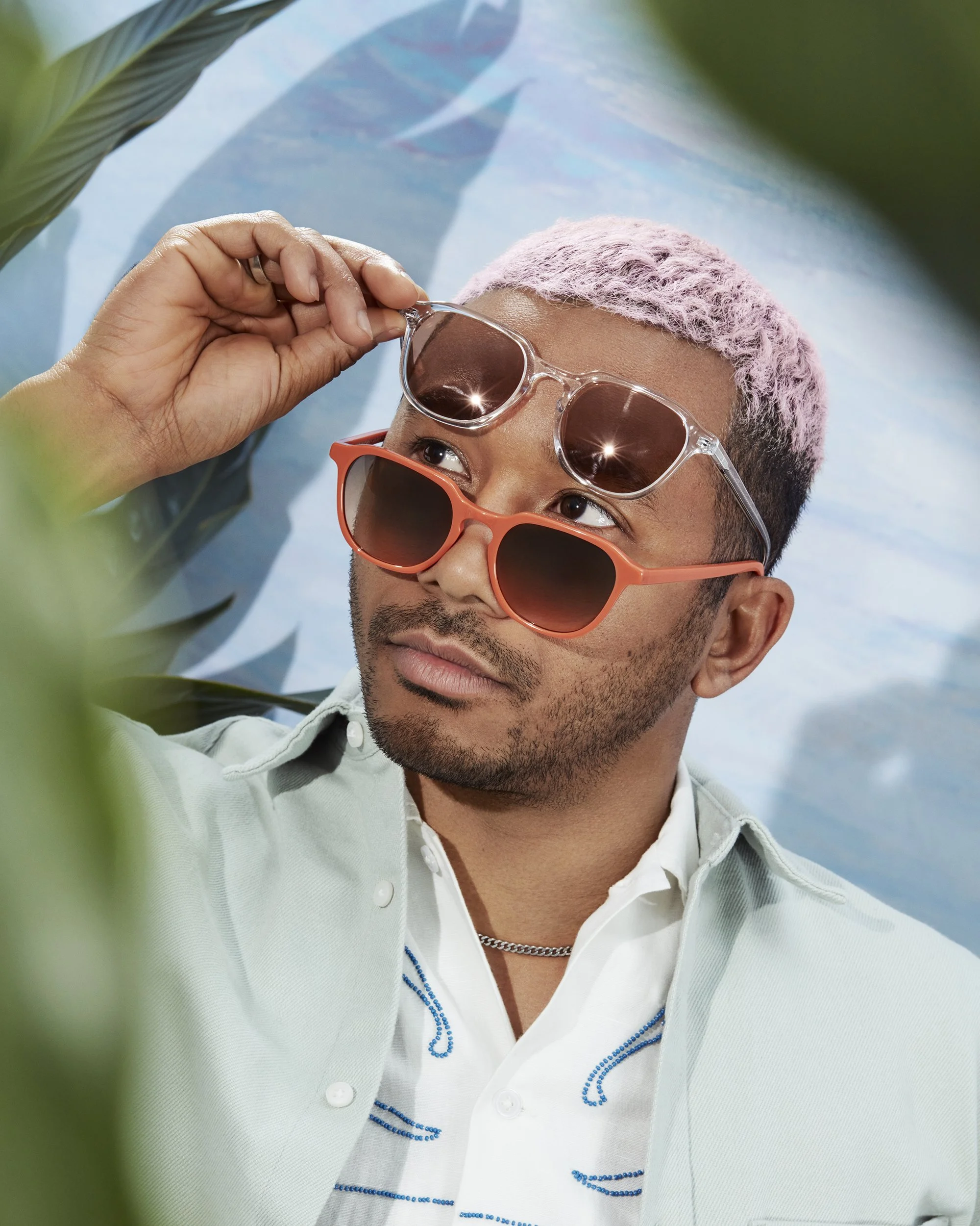 A man with pink hair and tan skin is wearing orange sunglasses propelled with black lenses, a white collared shirt with blue embroidery, a silver chain necklace, and is holding clear sunglasses with dark lenses up near his forehead, lying on a surface with leafy green plants around him.