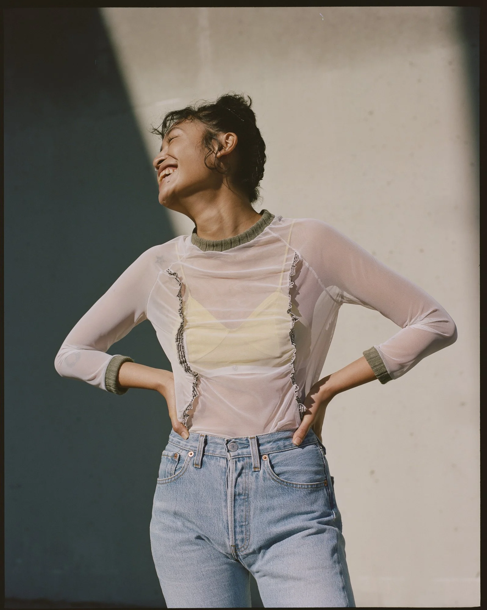 A young woman with short curly hair smiling, wearing a sheer long-sleeve top over a yellow camisole and high-waisted blue jeans, standing with her hands on her hips in front of a plain wall.