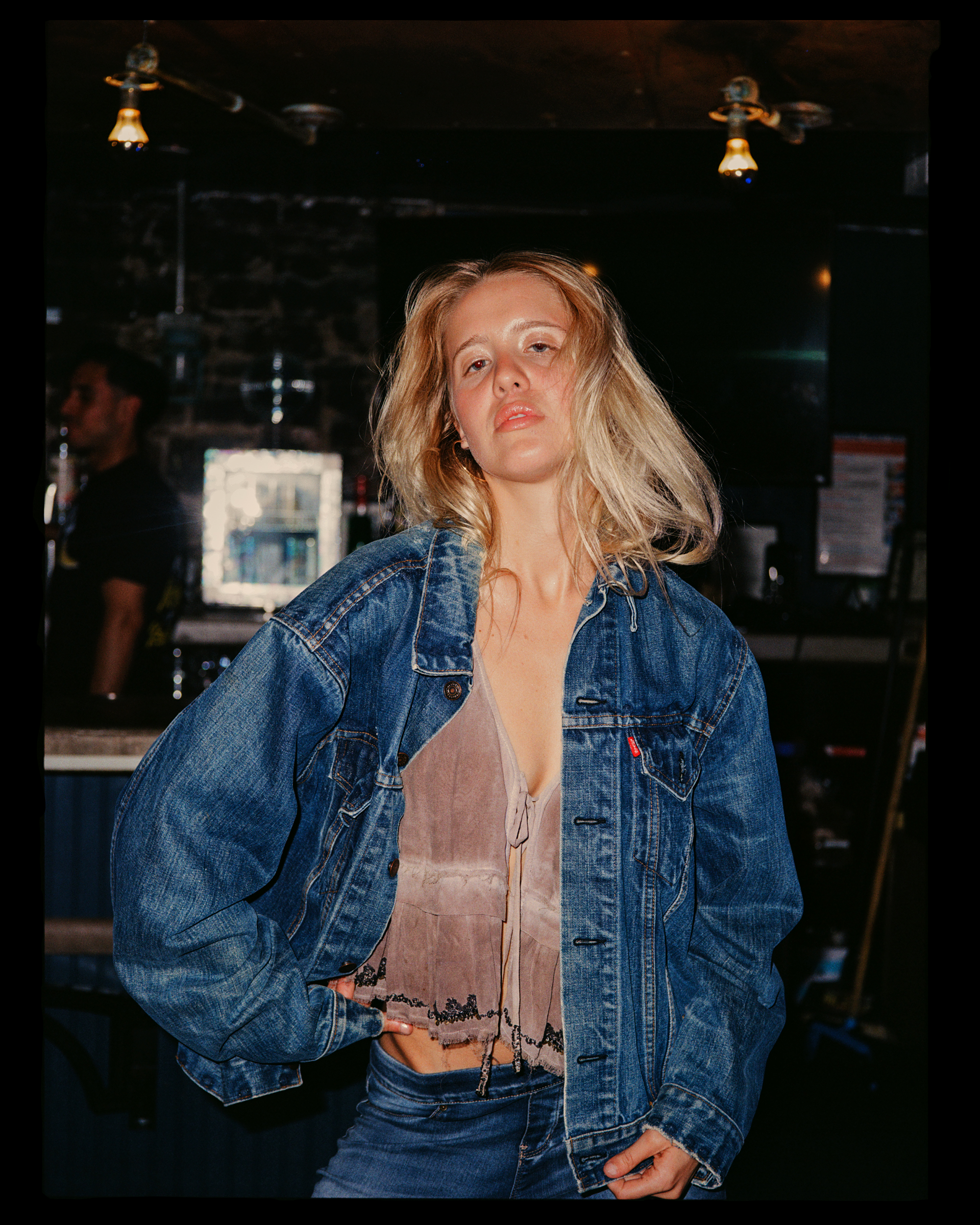 A woman with blonde hair wearing a denim jacket over a beige top with lace trim, standing in front of a dark background in a dimly lit room.