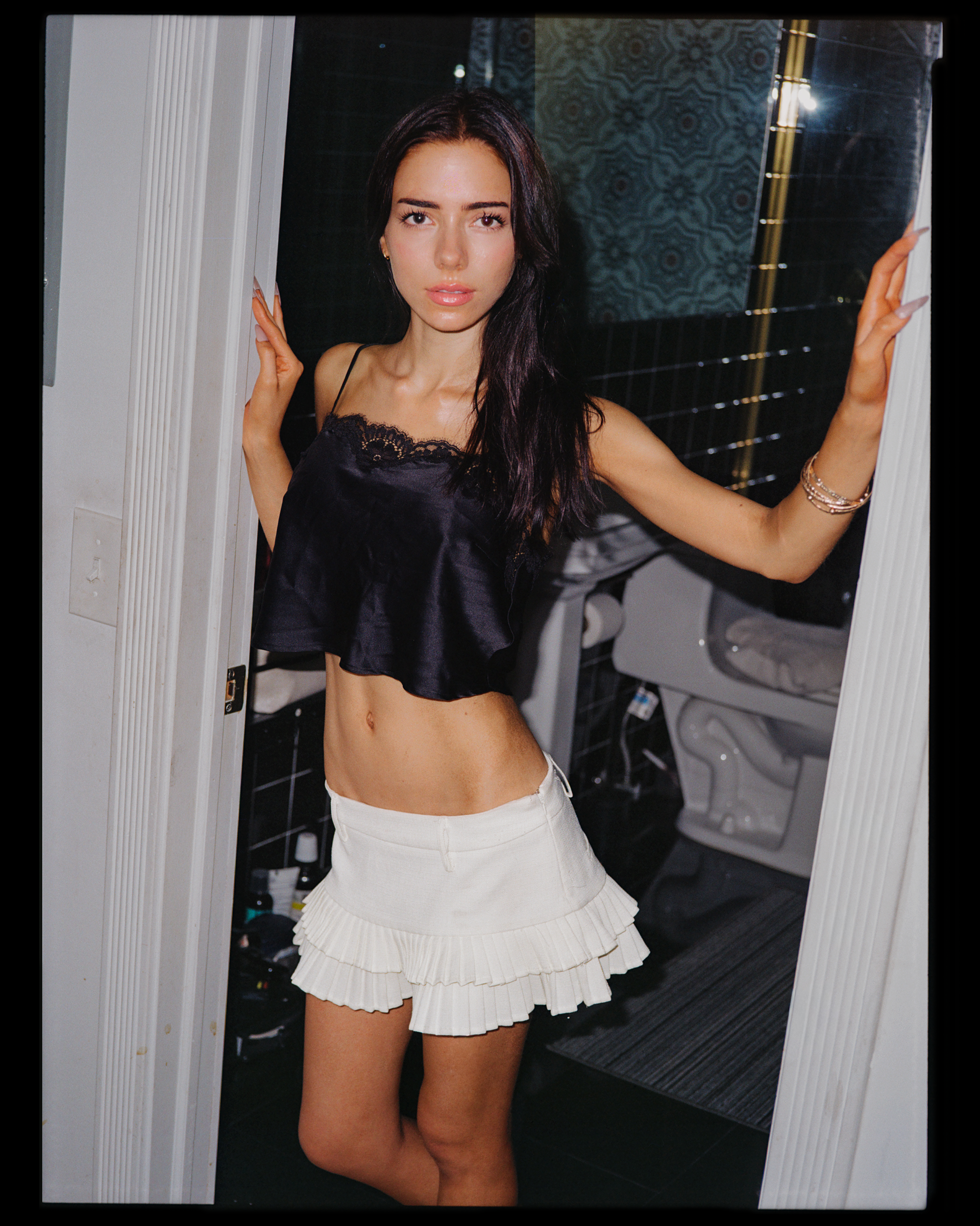 A young woman with long dark hair taking a mirror selfie in a bathroom, wearing a black satin camisole and a white ruffled skirt.