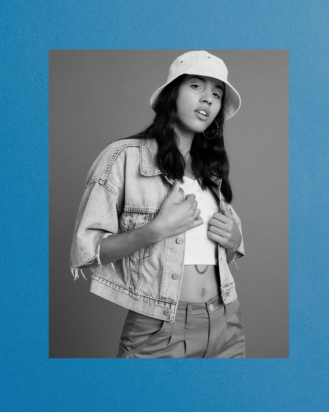A young woman poses in a studio with a gray background, wearing a white bucket hat, a distressed denim jacket, a white crop top, and high-waisted pants. She has long dark hair, hoop earrings, and slightly parted lips.