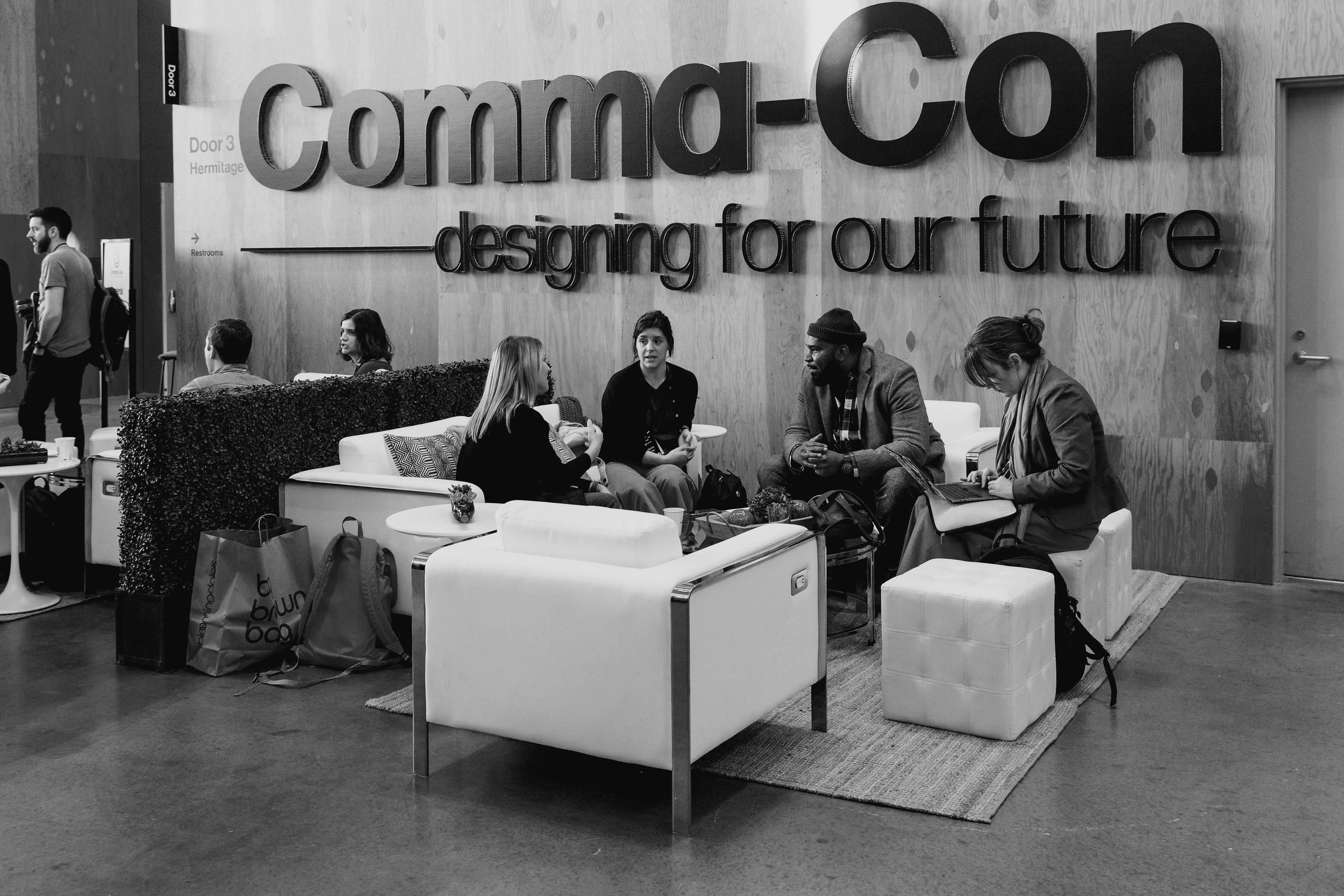 A modern lobby or waiting area with a large wall sign reading 'Commod-Corn - designing for our future.' Several people are seated on white sofas, engaged in conversation, with bags and laptops nearby. A man walks past in the background.