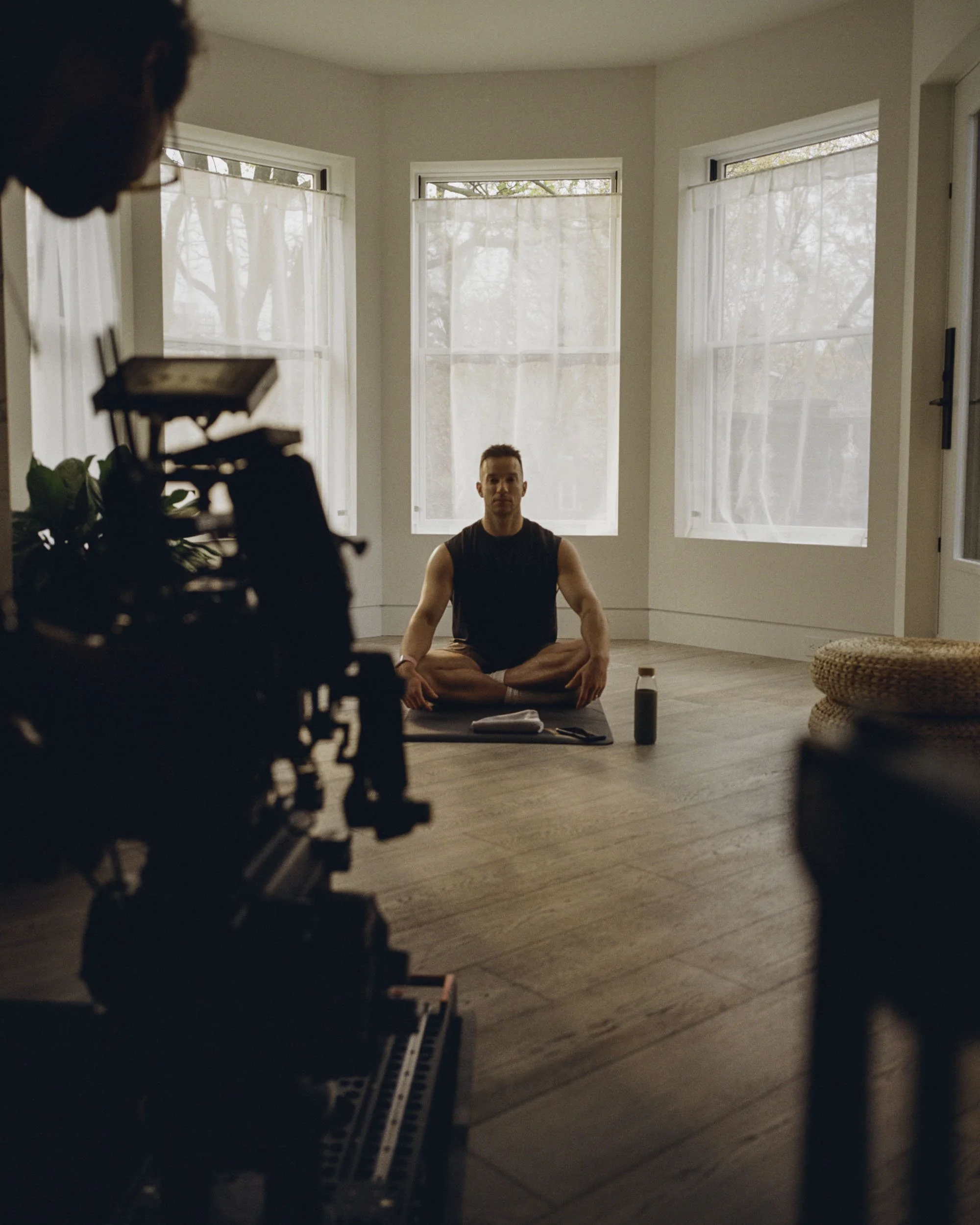 A man practicing yoga in a living room with large windows and light-colored walls, sitting cross-legged on a yoga mat.