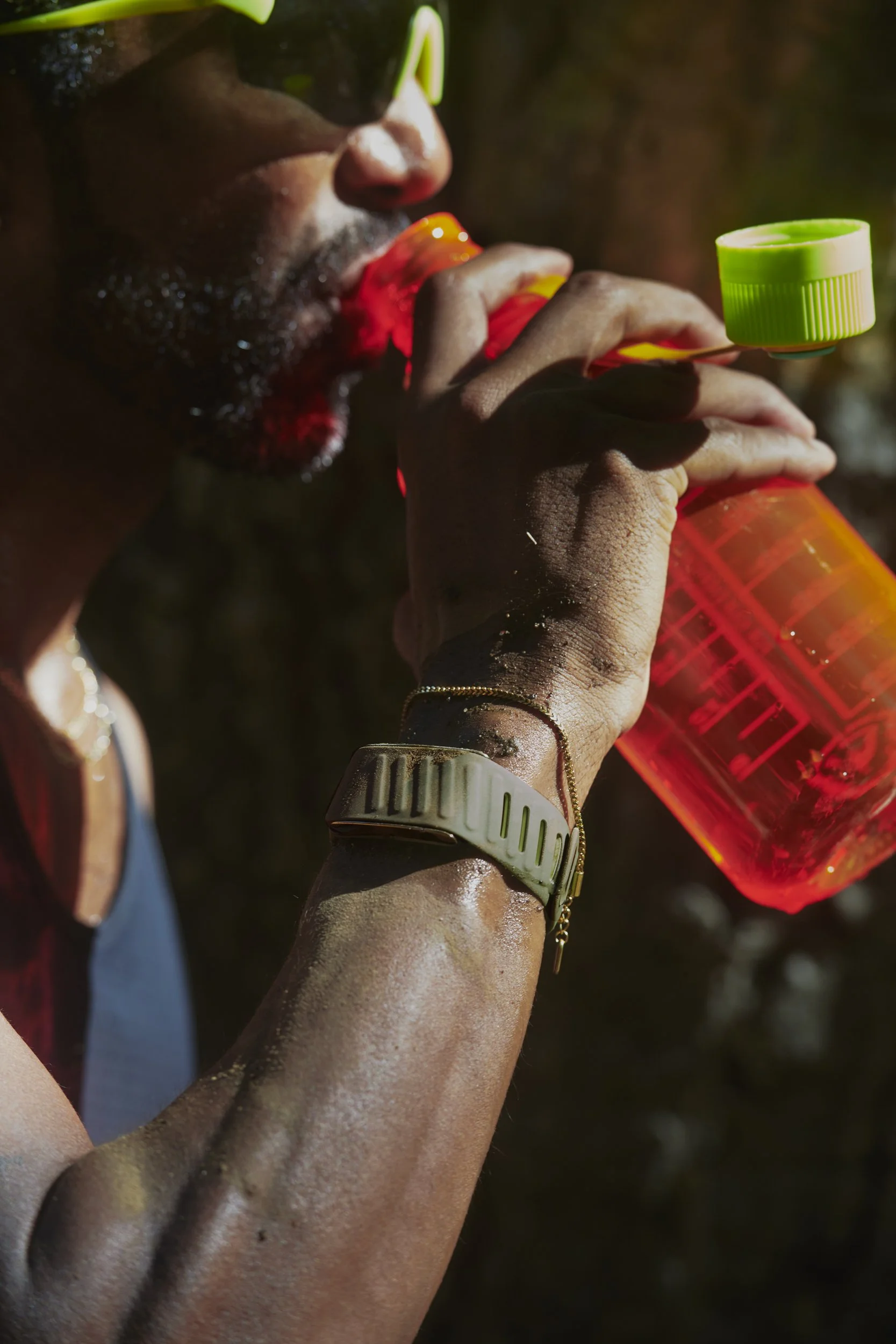 A person with dark skin drinking from a bright orange water bottle with a green cap, wearing sunglasses, a beige watch, and gold jewelry. The person is outdoors in sunlight.