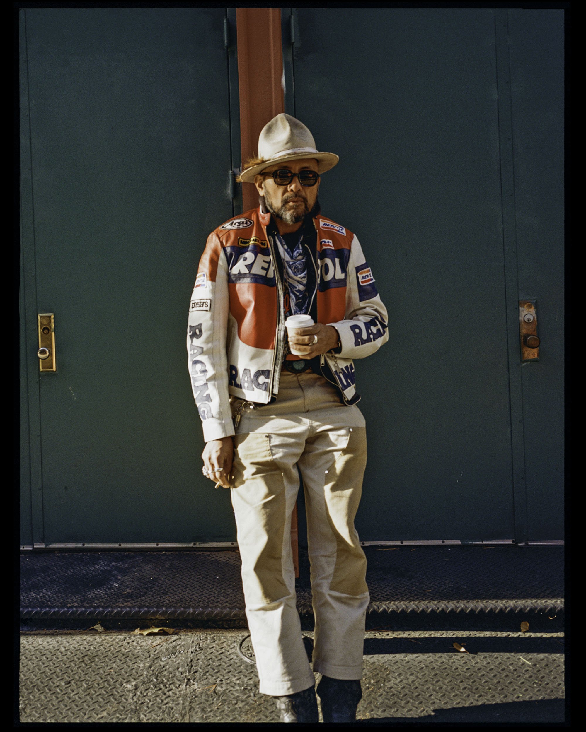 A man wearing a beige hat, sunglasses, a racing jacket, and beige pants is standing in front of a green door, holding a coffee cup.