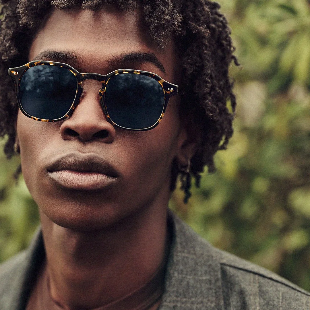 Close-up of a person with dark skin wearing tortoiseshell sunglasses and a gray jacket, outdoors with blurred green foliage in the background.