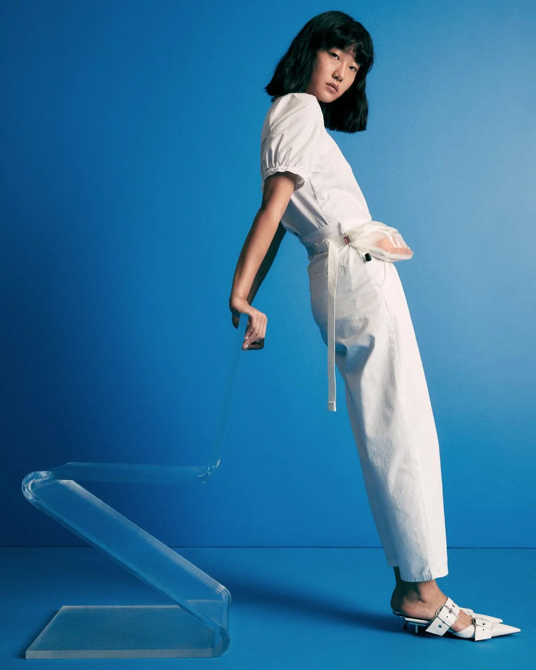 A woman with black hair in a white outfit, leaning against a clear plastic chair with a blue background.